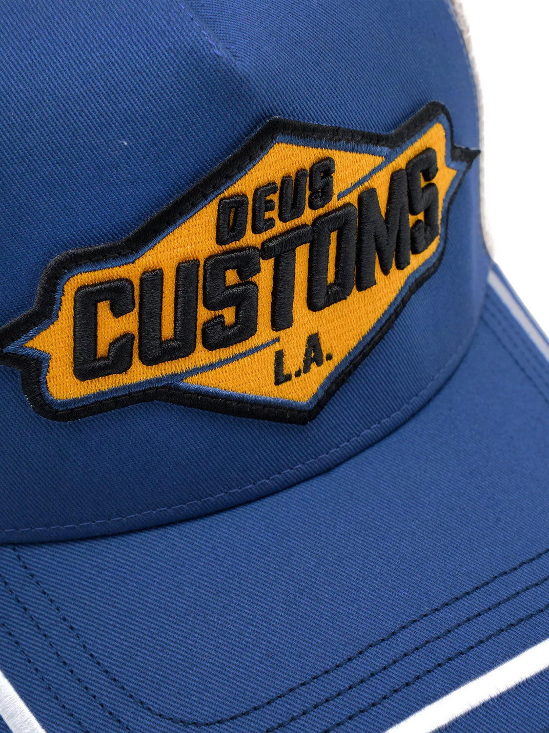 Close-up of the Deus Bills Trucker hat in blue, featuring a yellow and black “DEUS CUSTOMS L.A.” embroidered patch on the front, white stitching on the brim, and an adjustable snapback closure.