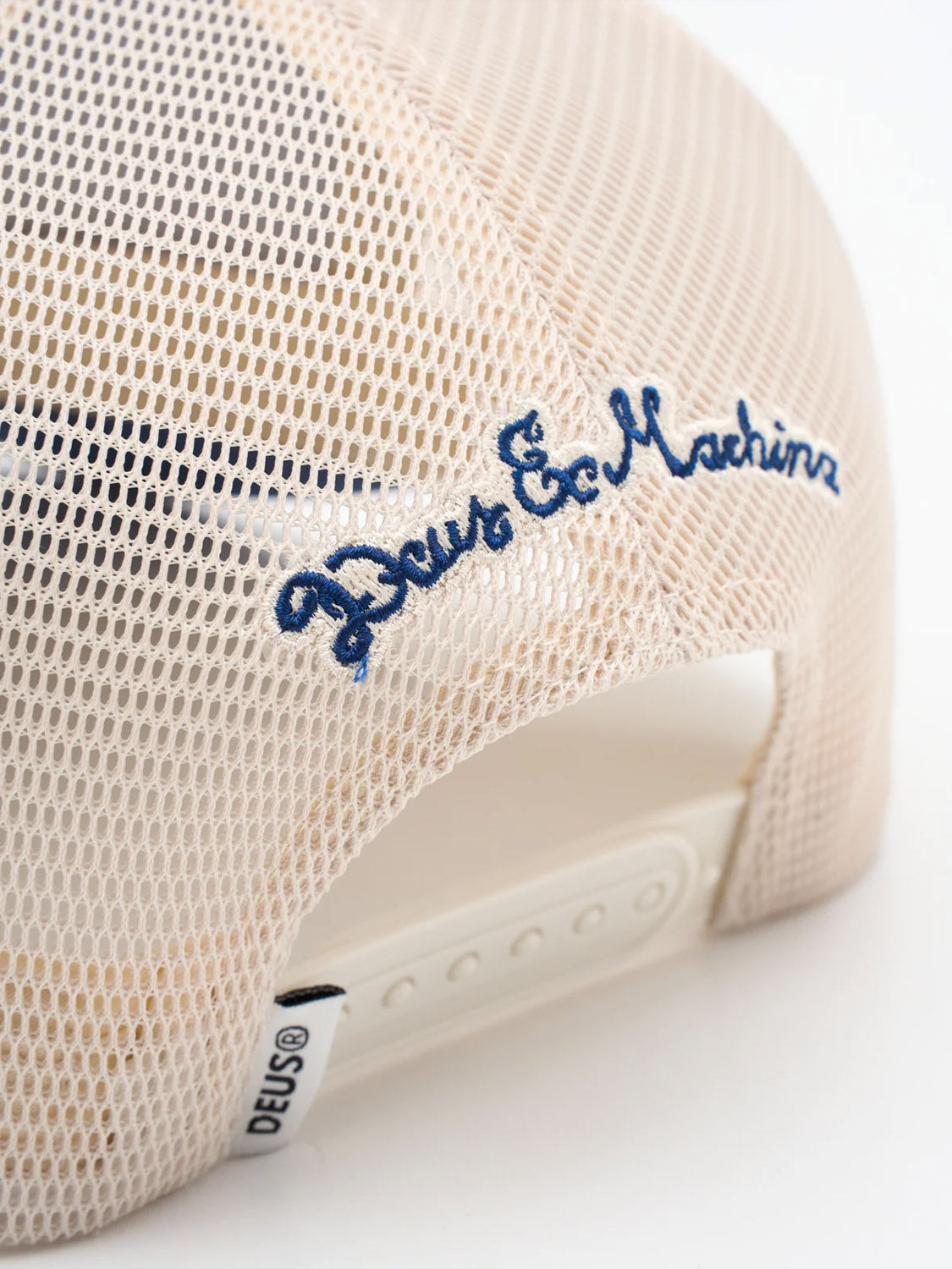 Close-up of the back of the Bills Trucker by Deus, a beige mesh hat with blue embroidered Deus Ex Machina artwork, an adjustable white snapback, and a signature white DEUS® tag near the strap.