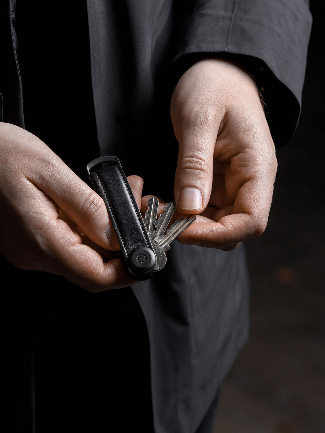 A person in a dark coat holds the sleek Orbitkeys Key Organizer Leather, displaying several metal keys fanned out in their hands against a dark, blurred background.