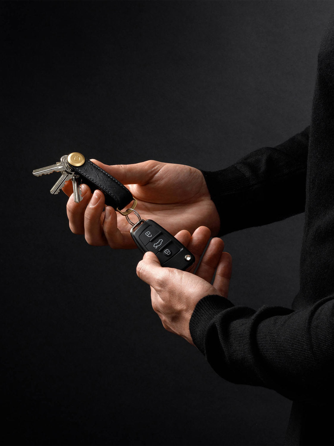 A person in a black long-sleeve shirt holds keys and an Orbitkeys Key Organizer Saffiano Leather with a black car key fob against a dark background.