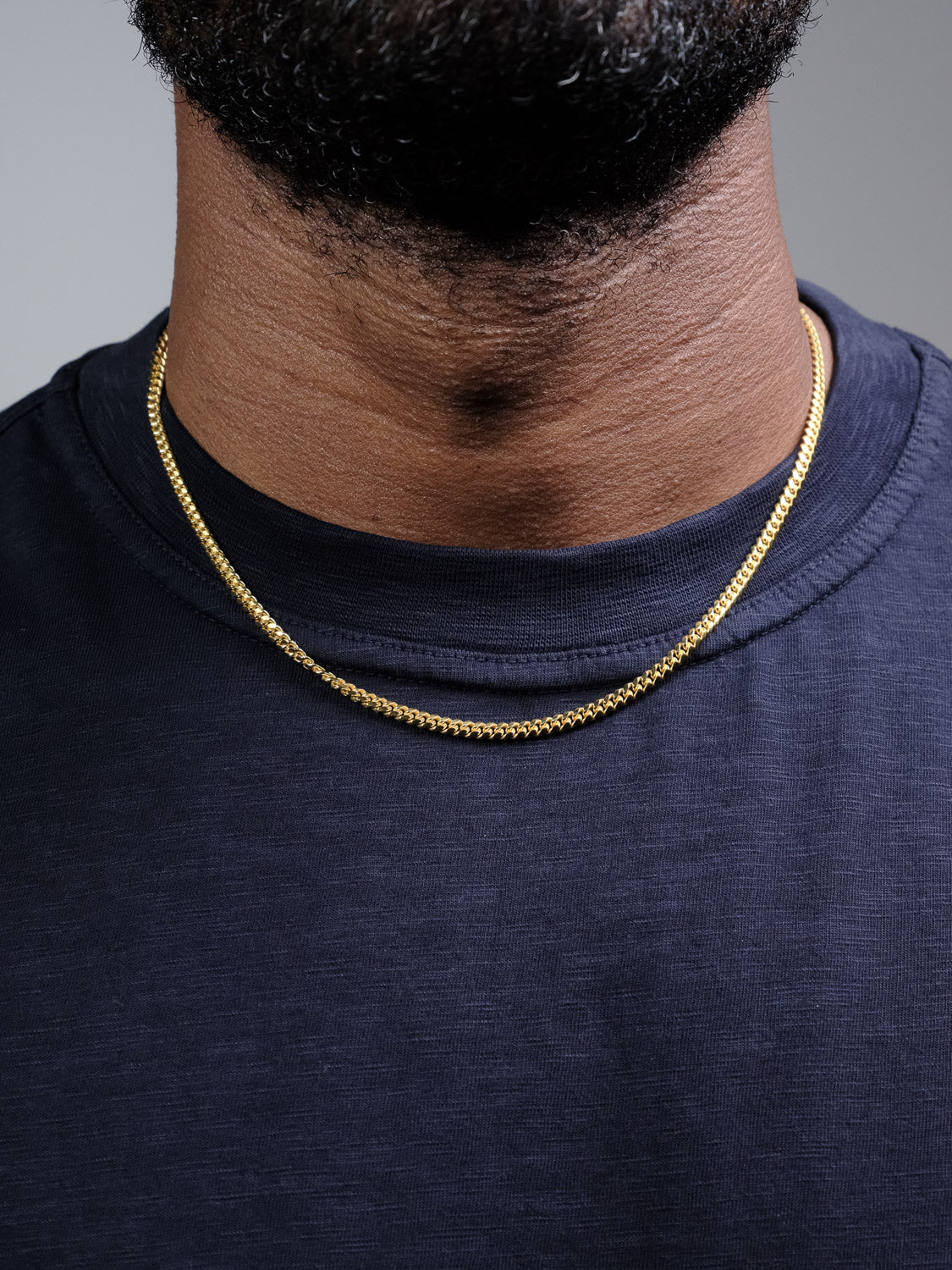 A close-up shows a person wearing the Blaze 80 Miami Cuban Necklace by ID Menswear with a navy blue crew-neck shirt. Only the lower face and neck are visible; the rest is out of frame.