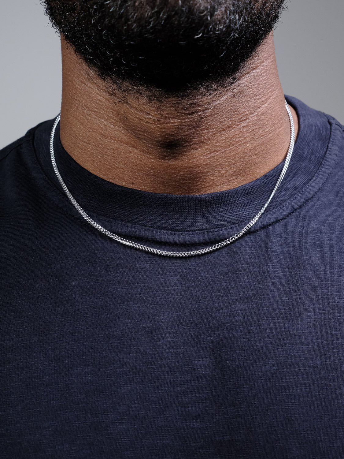 A close-up shows a person wearing a dark blue T-shirt and the Blaze 100 Miami Cuban Necklace by ID Menswear. The focus is on the neck, collar, and jawline, without revealing the face.
