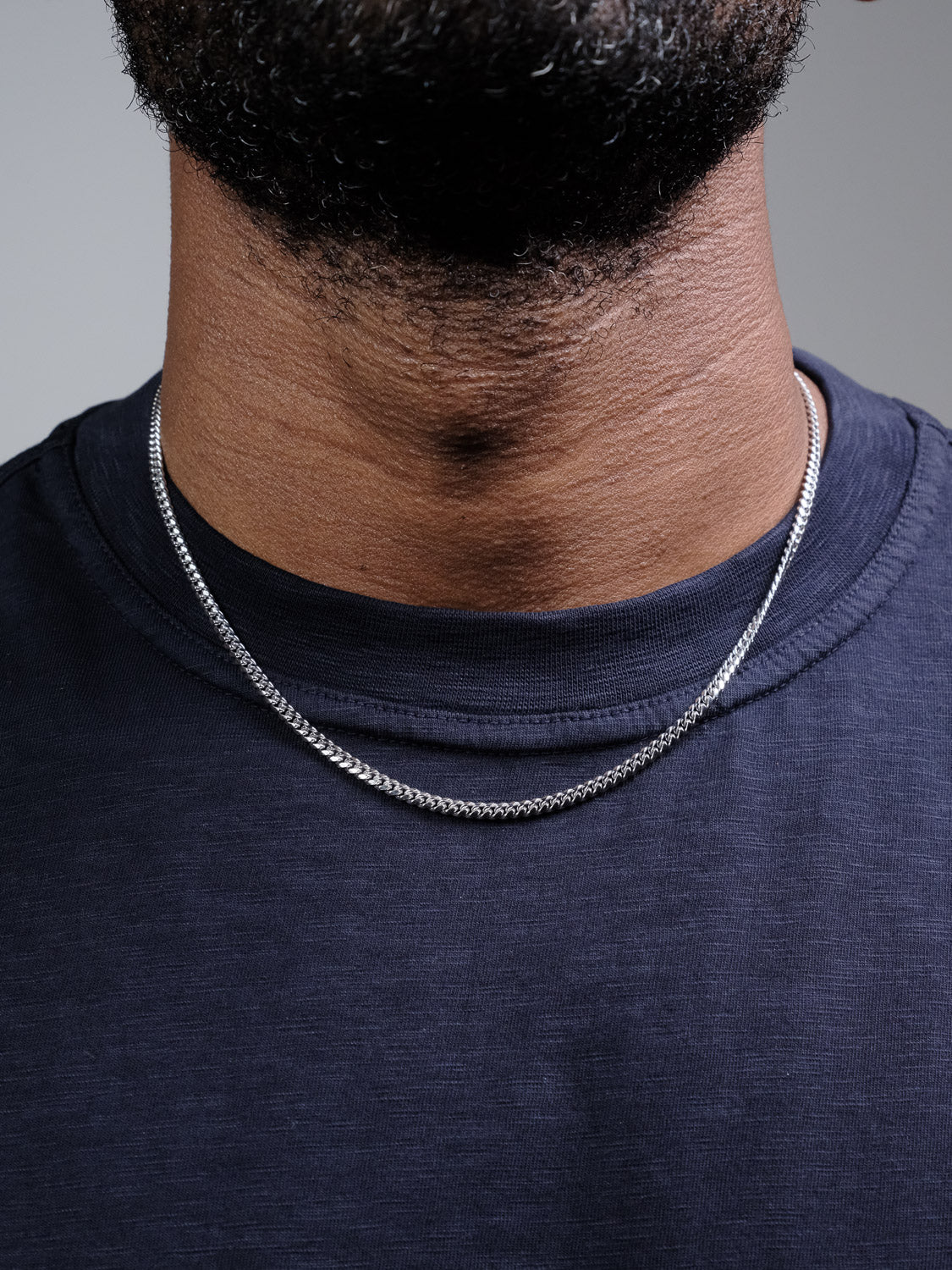 A close-up of a person’s neck wearing the Blaze 80 Miami Cuban Necklace by ID Menswear, made from 925 Sterling Silver, paired with a navy blue crew-neck shirt and part of their beard visible.