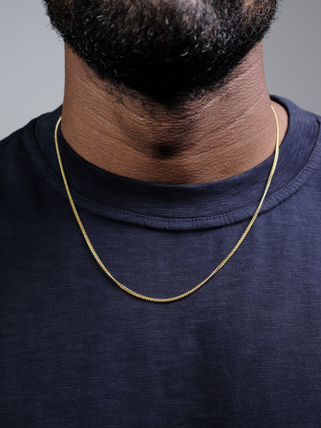 A close-up of a person's neck and upper chest shows them wearing the ID Menswear Blaze 60 Miami Cuban Necklace with a dark navy blue shirt. The person's face is not visible.