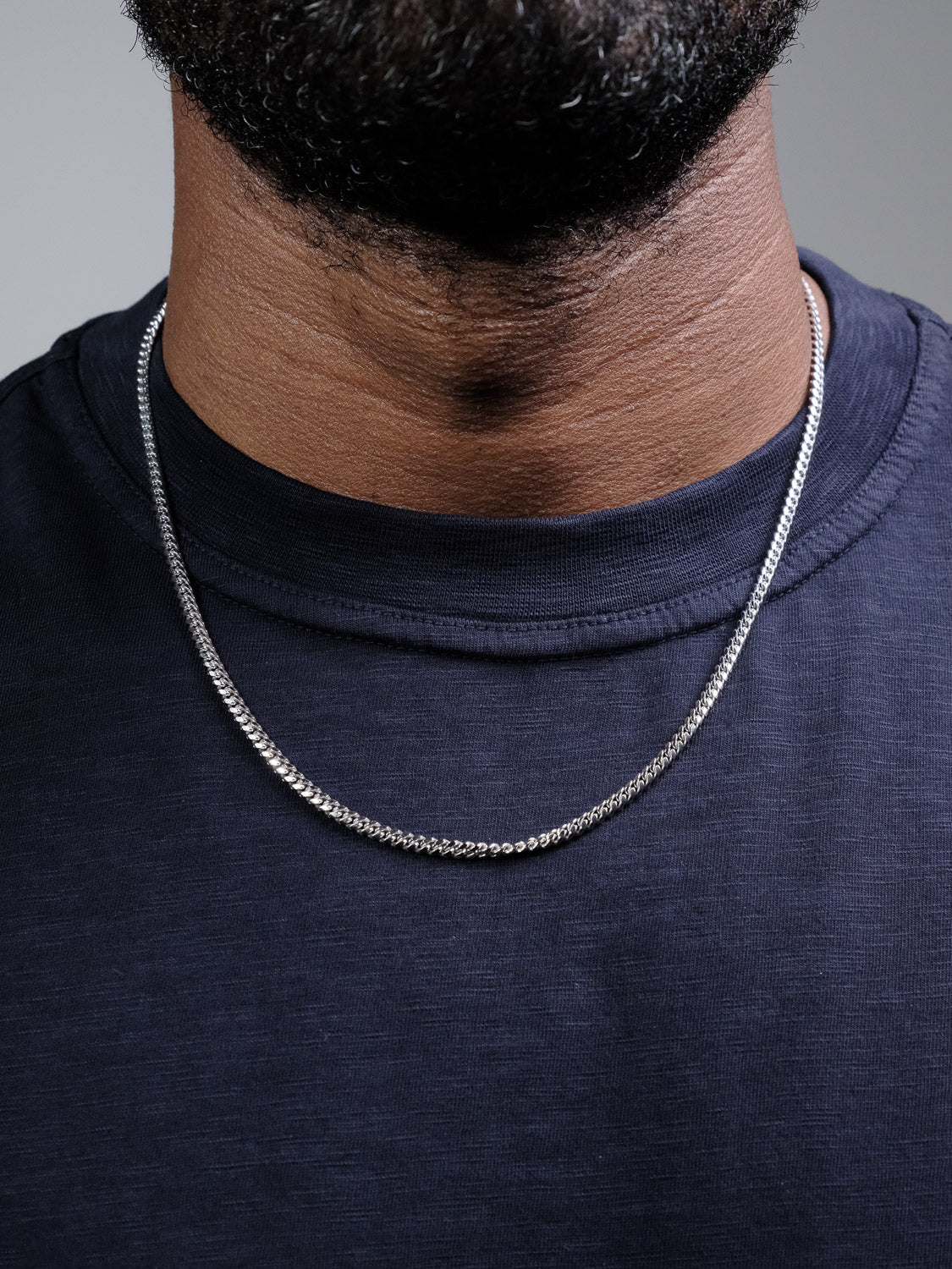 A close-up of a person wearing a dark crew-neck shirt and the Blaze 80 Miami Cuban Necklace by ID Menswear, a thin 925 Sterling Silver chain. The person's lower face, beard, and neck are visible.
