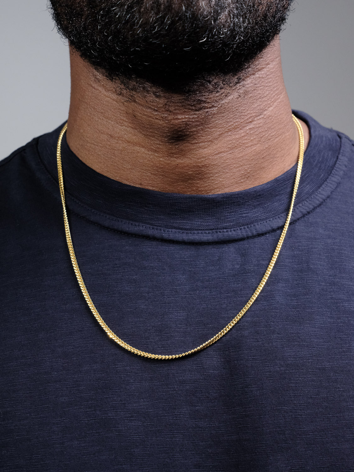 A close-up shows a person wearing a dark blue shirt and the Blaze 100 Miami Cuban Necklace by ID Menswear, with only the lower half of their face and neck visible.
