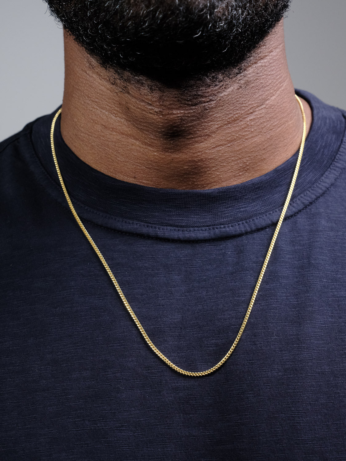 A close-up shows a person wearing the Blaze 60 Necklace by ID Menswear, a 3mm gold-plated chain over a dark navy blue crew-neck shirt, with only the lower face, neck, and upper chest visible.