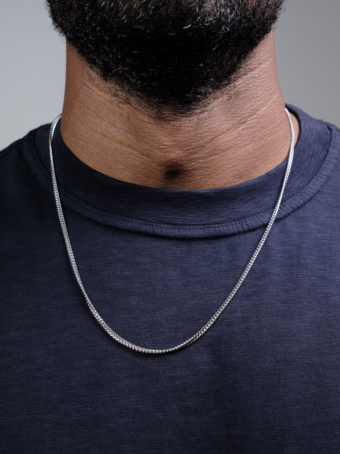 A close-up of a person wearing the Blaze 100 Miami Cuban Necklace by ID Menswear with a dark navy-blue t-shirt, showing only the lower half of their face and neck.