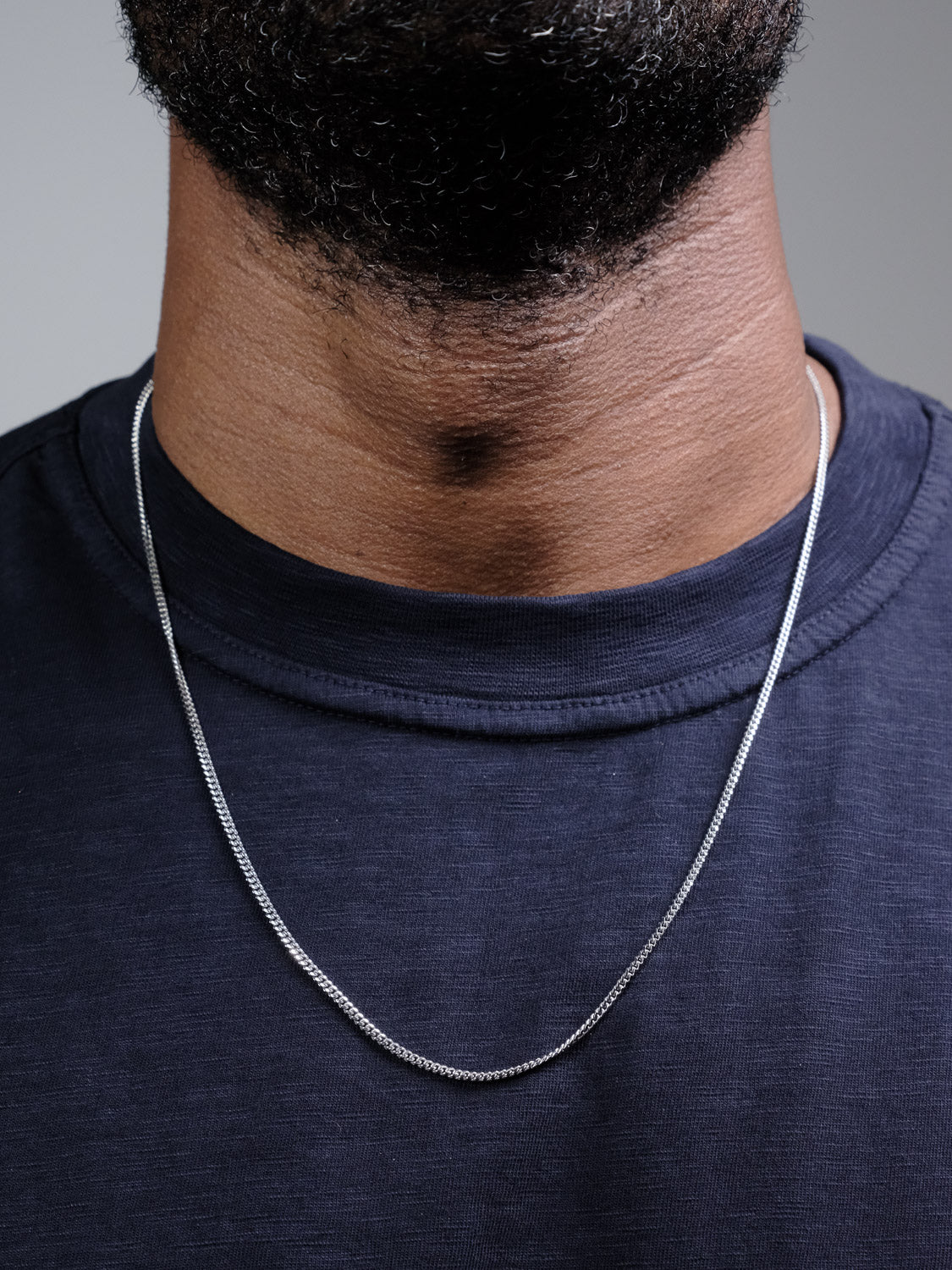A close-up of a person's neck and upper chest shows them wearing the Blaze 60 Necklace by ID Menswear, a thin sterling silver chain, over a navy blue crew-neck shirt. The lower beard and neckline are also visible.