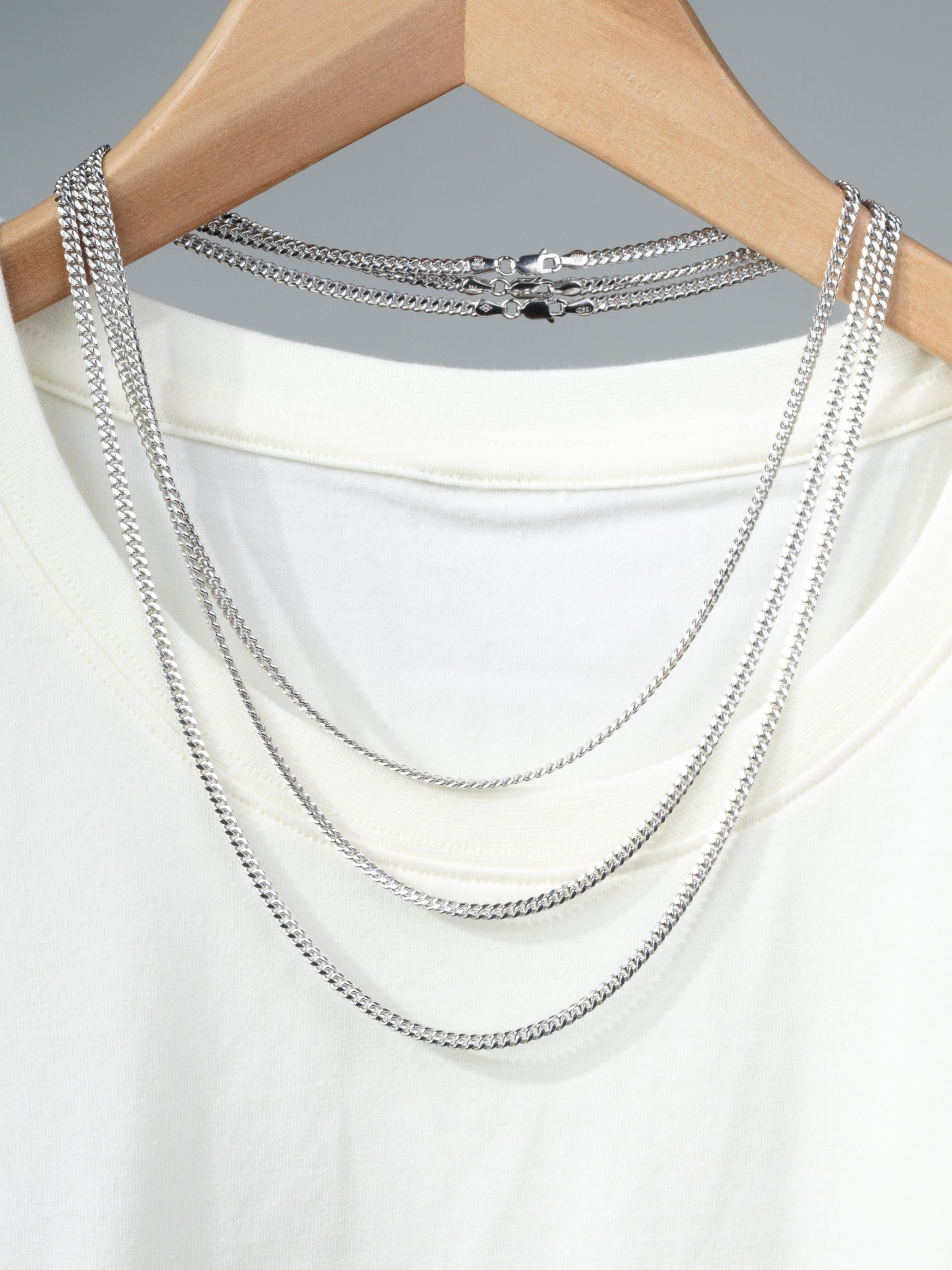 A white T-shirt on a wooden hanger showcases three layered silver chains, including the Blaze 100 Miami Cuban Necklace by ID Menswear, draped over the neckline against a plain gray background.