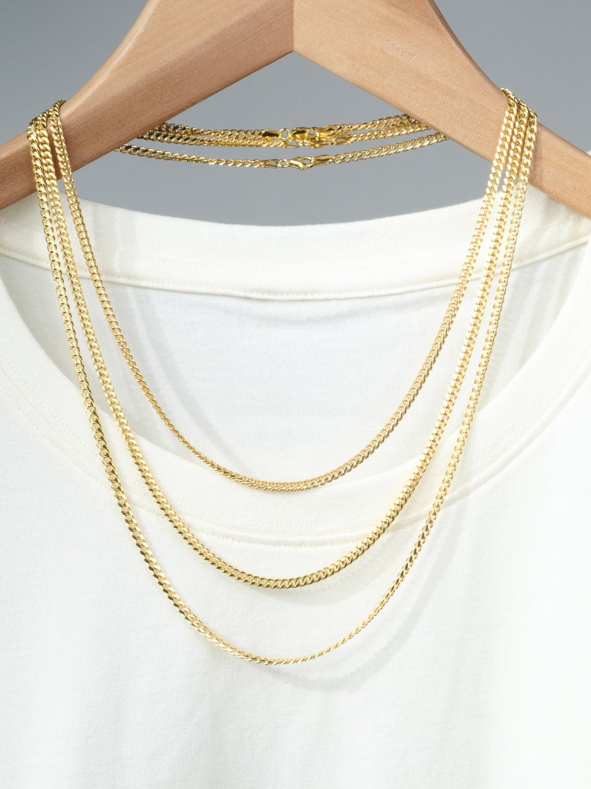 Three gold chain necklaces, featuring the Blaze 100 Miami Cuban Necklace by ID Menswear, draped over a wooden hanger with a plain white T-shirt against a simple gray background.
