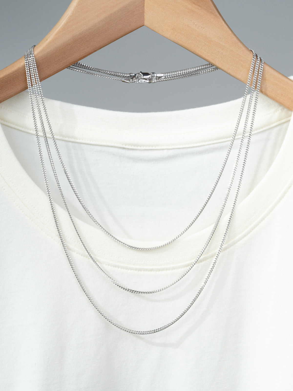 A white T-shirt on a wooden hanger features the Blaze 60 Miami Cuban Necklace by ID Menswear, crafted in layered 925 sterling silver and draped over the neckline, set against a plain gray background.