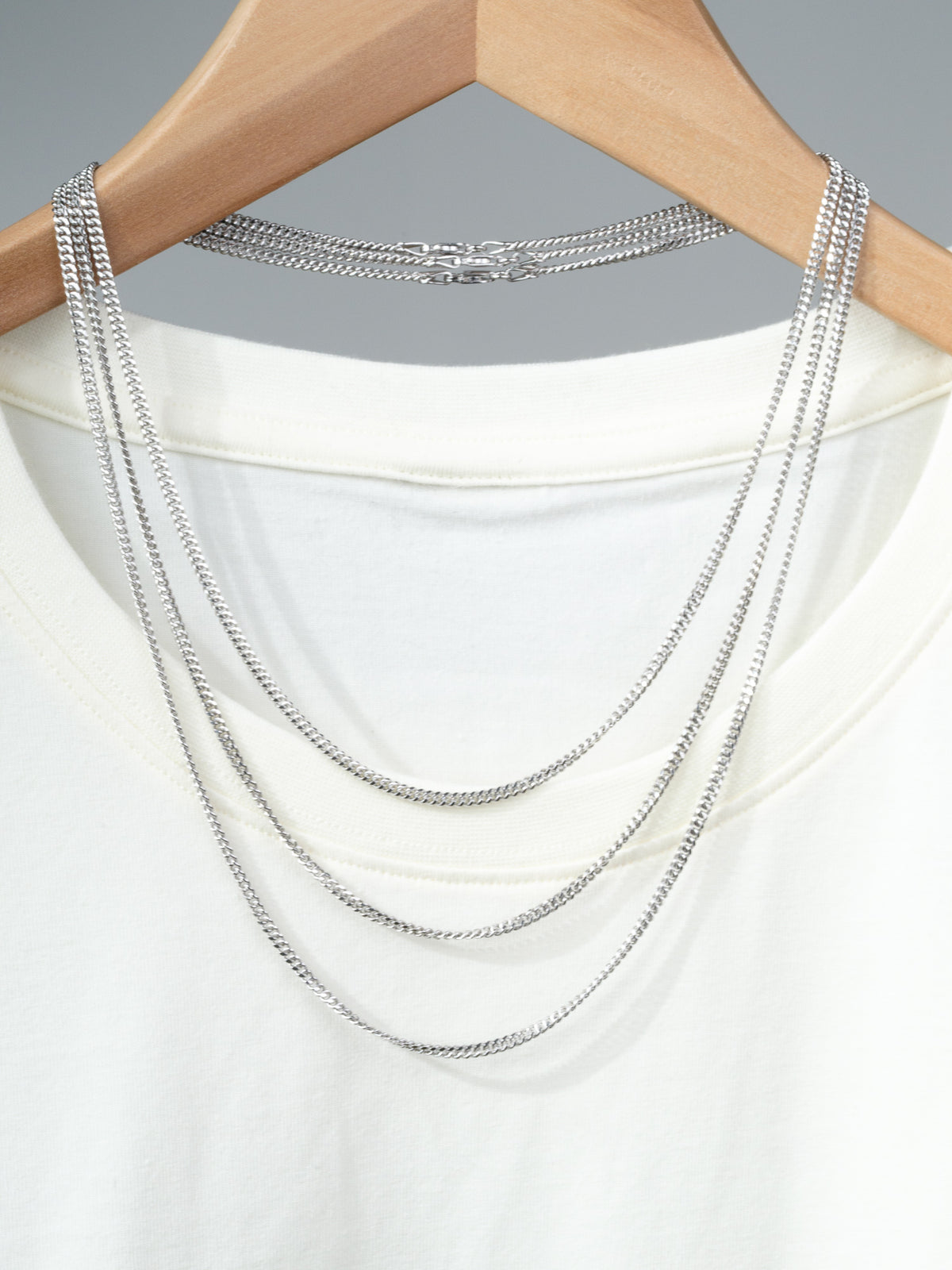 A white T-shirt on a wooden hanger displays three layered 925 Sterling Silver chains, including the Blaze 80 Miami Cuban Necklace by ID Menswear, draped over the neckline against a plain gray background.