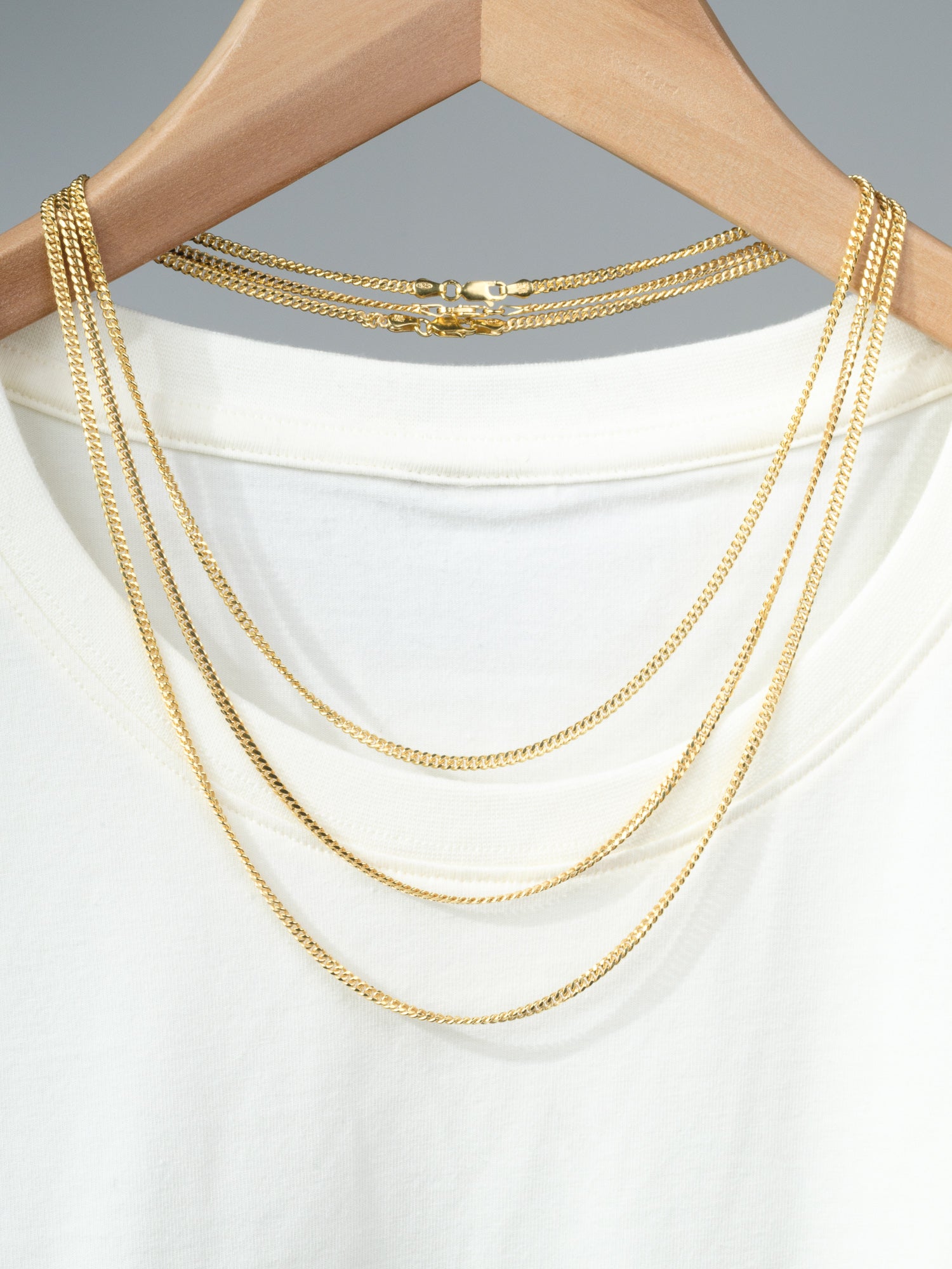 A white T-shirt on a wooden hanger with three layered gold-plated necklaces, including the Blaze 80 Miami Cuban Necklace by ID Menswear, draped over the neckline against a neutral background.