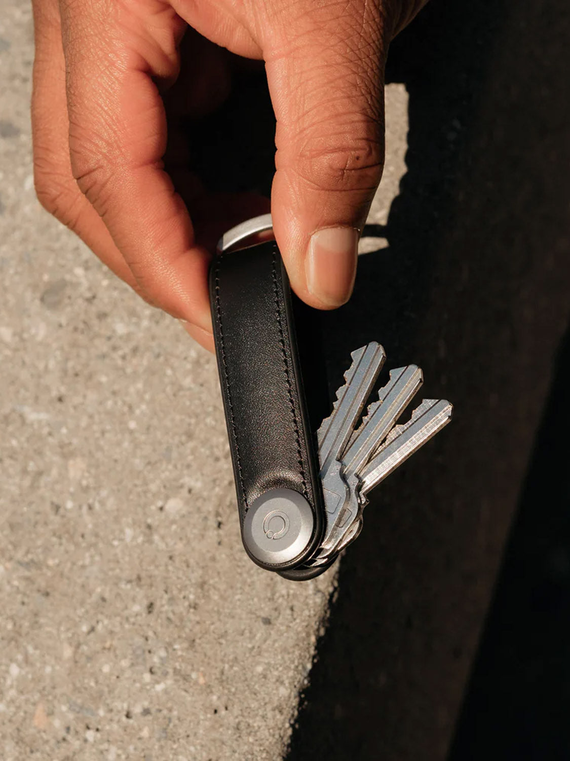 A hand holds the Orbitkeys Key Organizer Hybrid Leather in black, displaying its secure locking mechanism and neatly stacked metal keys, set against a rough concrete surface.