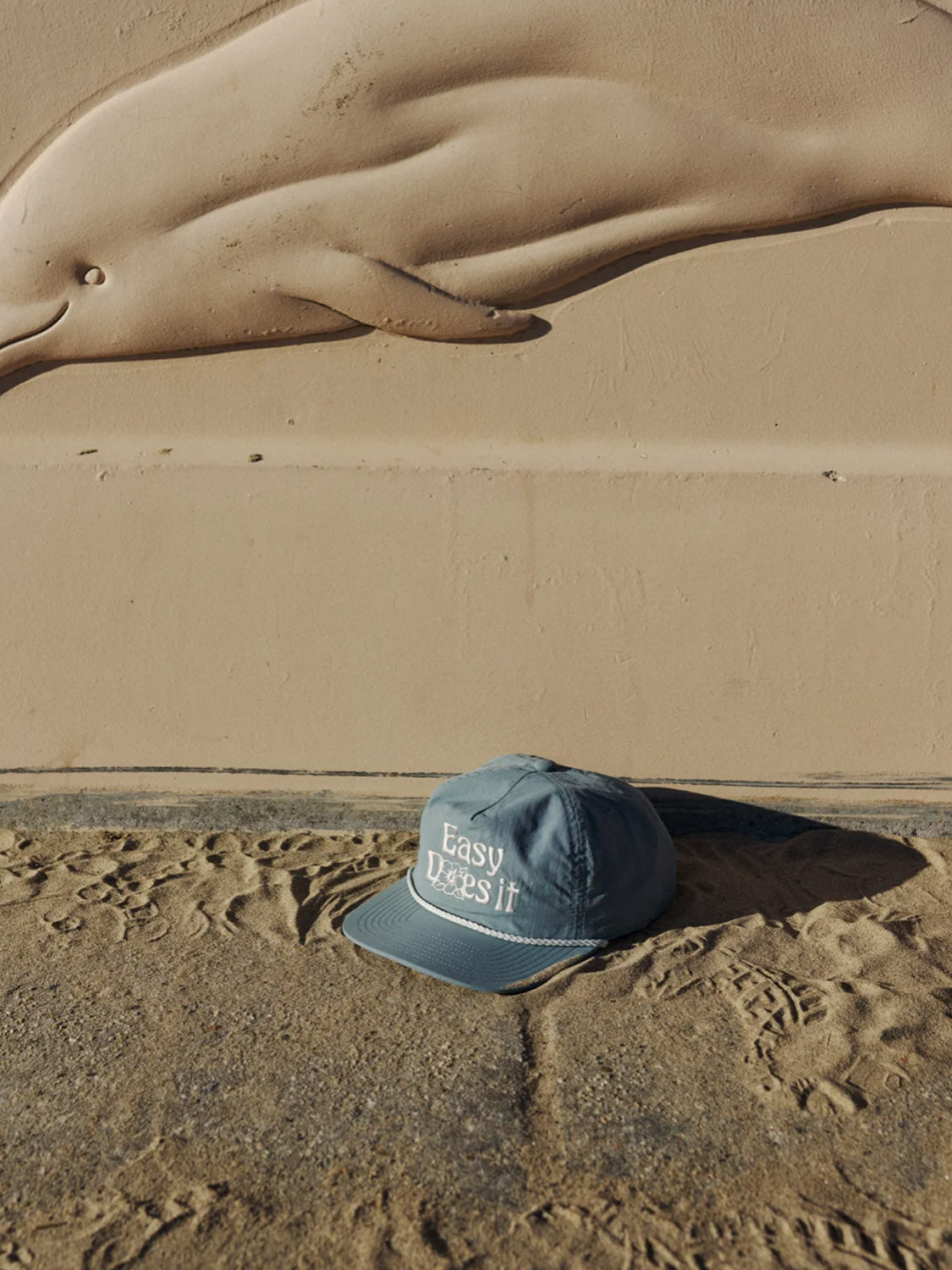 The Katin Bloom Hat, a blue 5-panel cap, rests on sand in front of a beige wall showcasing a raised dolphin design.