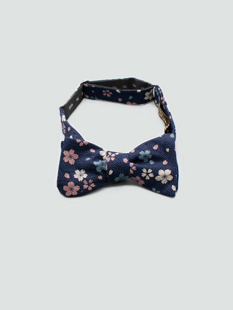 The ID Japanese Printed Blossom Bow Tie by ID Menswear is a navy blue cotton accessory with small pink and white flower prints, handmade in Brooklyn and shown against a light gray background.