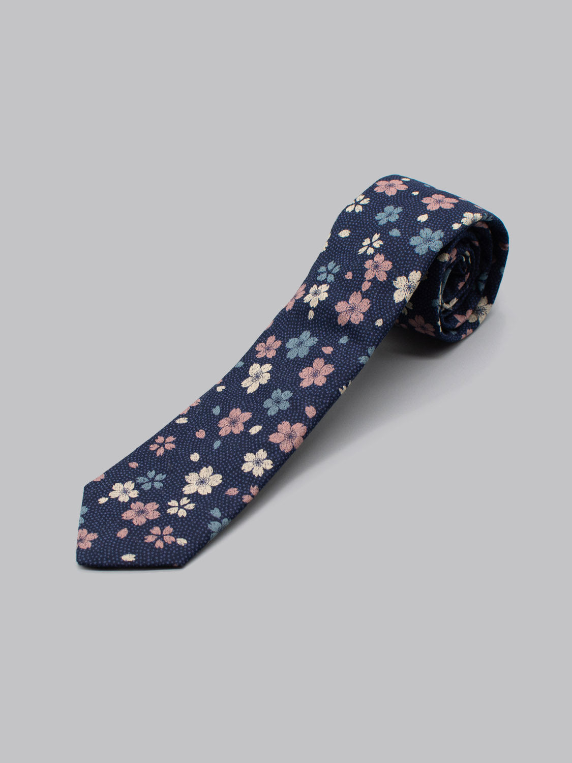 The Japanese Printed Blossom Tie by ID Menswear features a navy blue base with pink, white, and blue floral patterns. Handmade in Brooklyn from 100% cotton, it’s shown rolled at one end on a plain light gray background.
