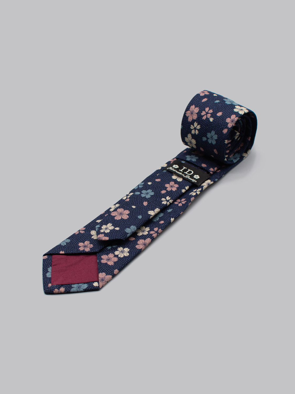 The Japanese Printed Blossom Tie by ID Menswear is handmade in Brooklyn from 100% cotton, featuring pink, white, and light blue floral print on navy. Partially unrolled on a gray background, it has a maroon back accent and visible fabric label.