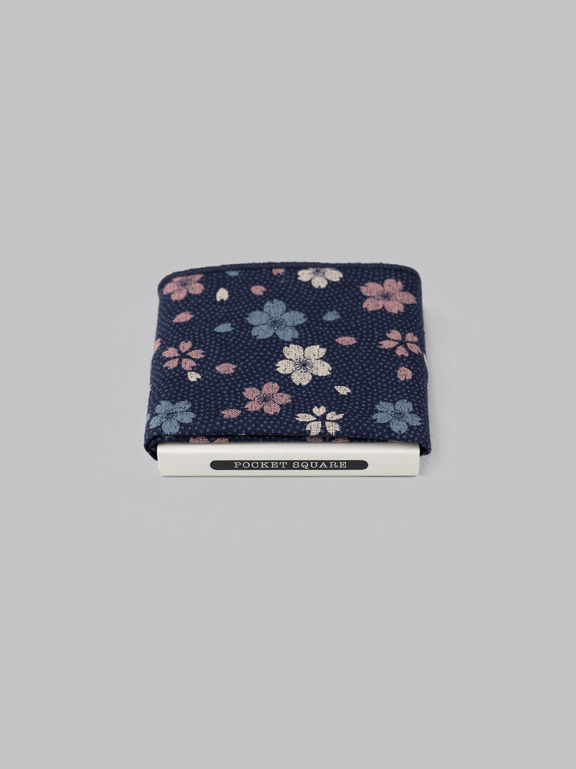 The Blossom Pocket Square by ID Menswear, handmade in Brooklyn, features a navy blue Japanese print with pink, white, and light blue flowers. Its shown folded against a plain gray background.