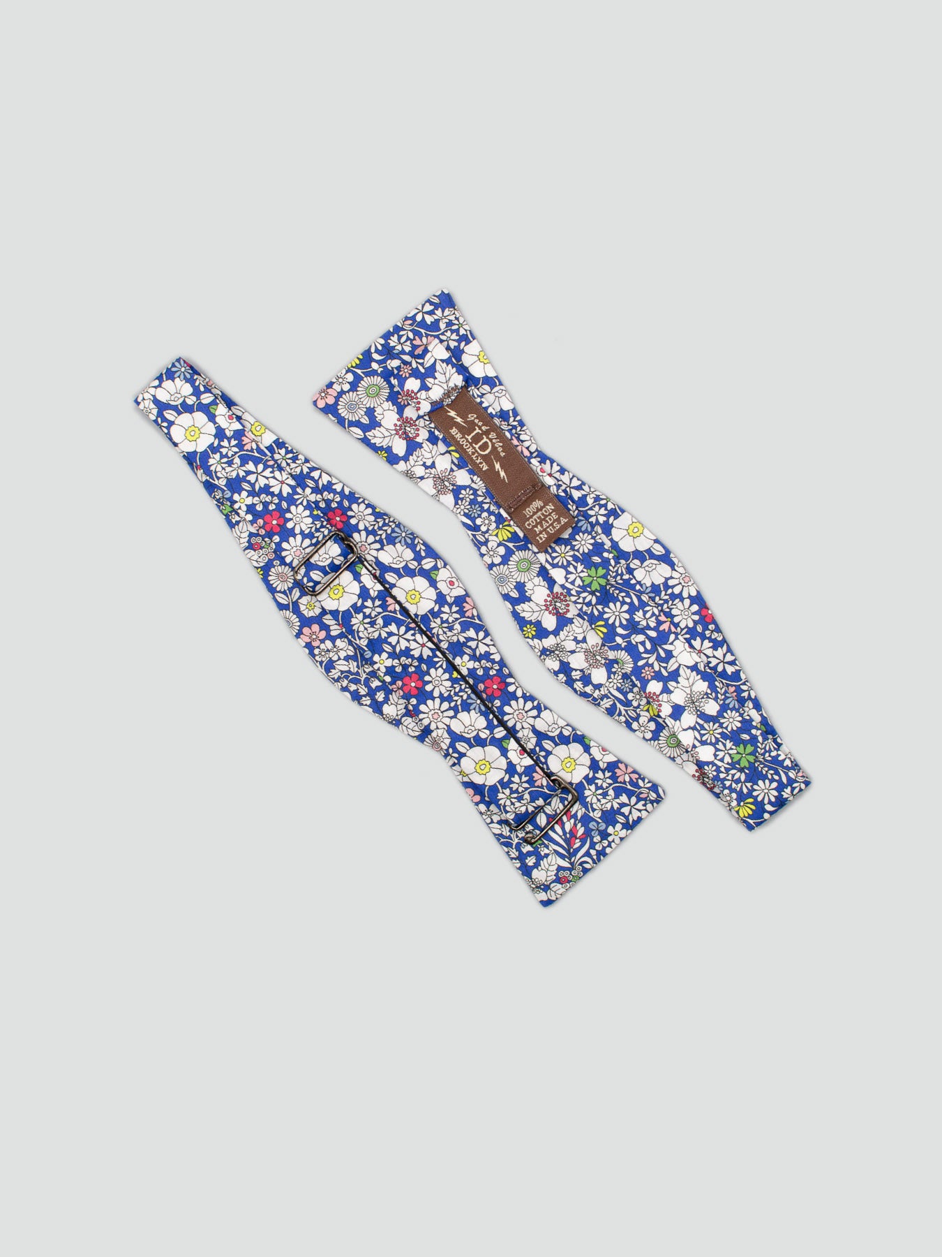 Two Liberty of London Tana Lawn™ Blue Lawn bow ties by ID Menswear, featuring a white, yellow, and green floral print—one shows the front and the other displays a brown label plus metal clasp—set on a light grey background.