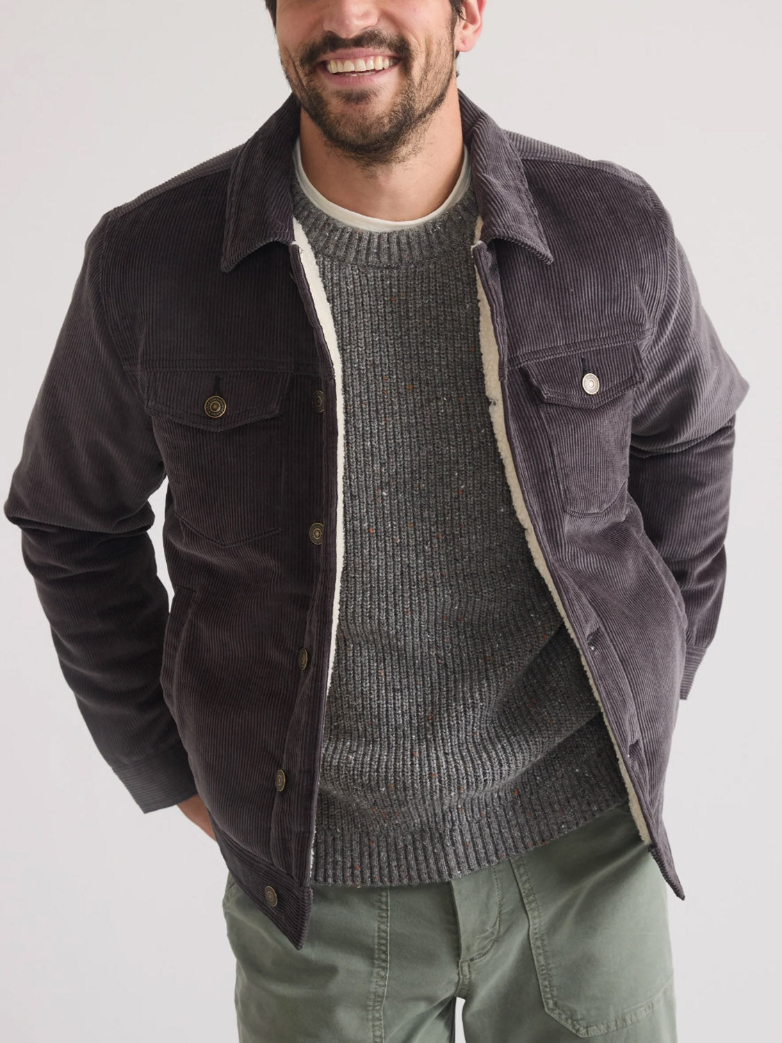 A man stands smiling with his hands in his pockets, wearing the Marine Layer Boise Cord Trucker jacket over a gray knit sweater and green pants, set against a plain light background.