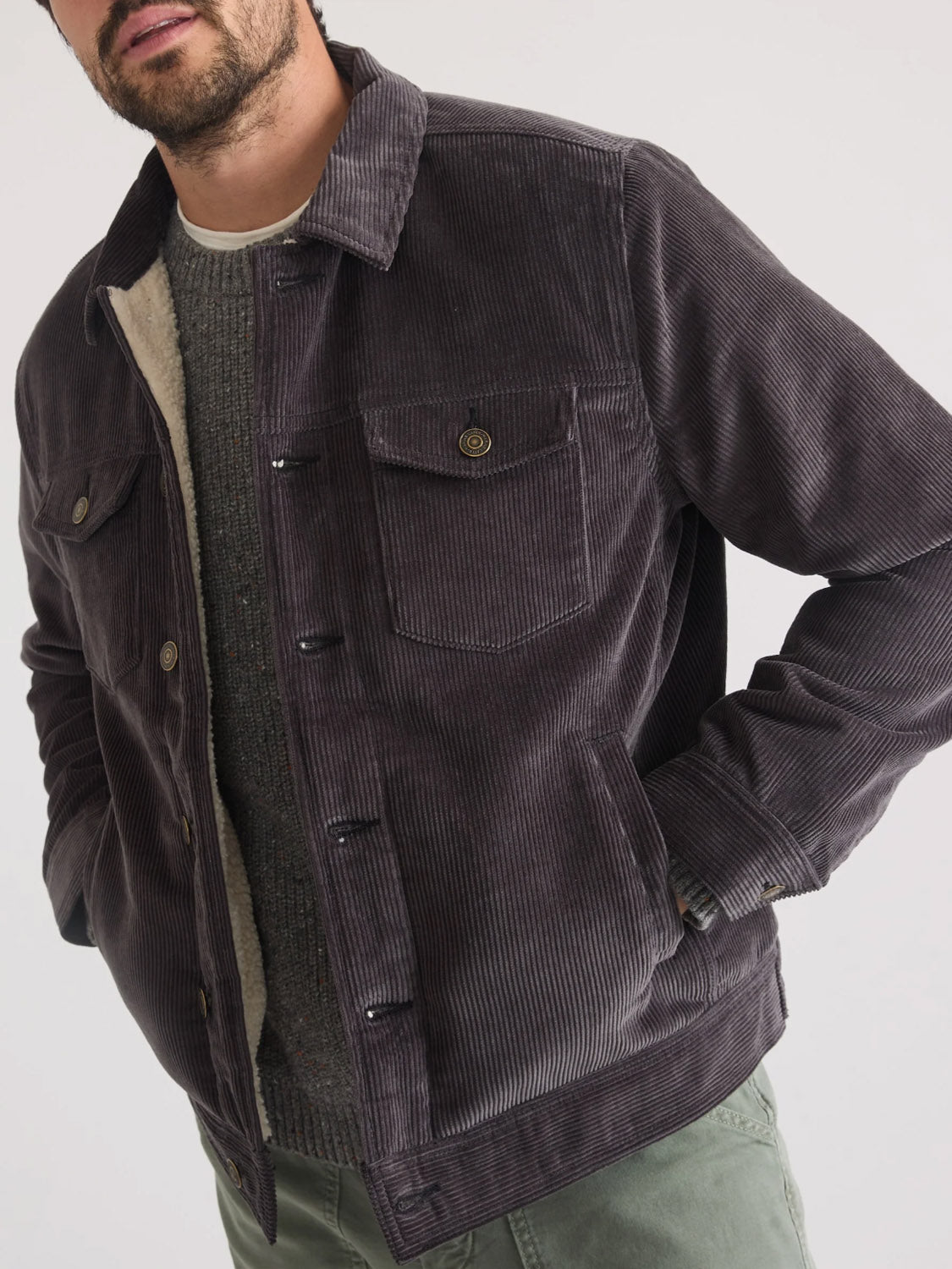 A man wearing the Marine Layer Boise Cord Trucker, a men's classic fit dark gray corduroy jacket with a buttoned front, layered over a textured sweater and green pants, stands with one hand in his pocket against a plain background.