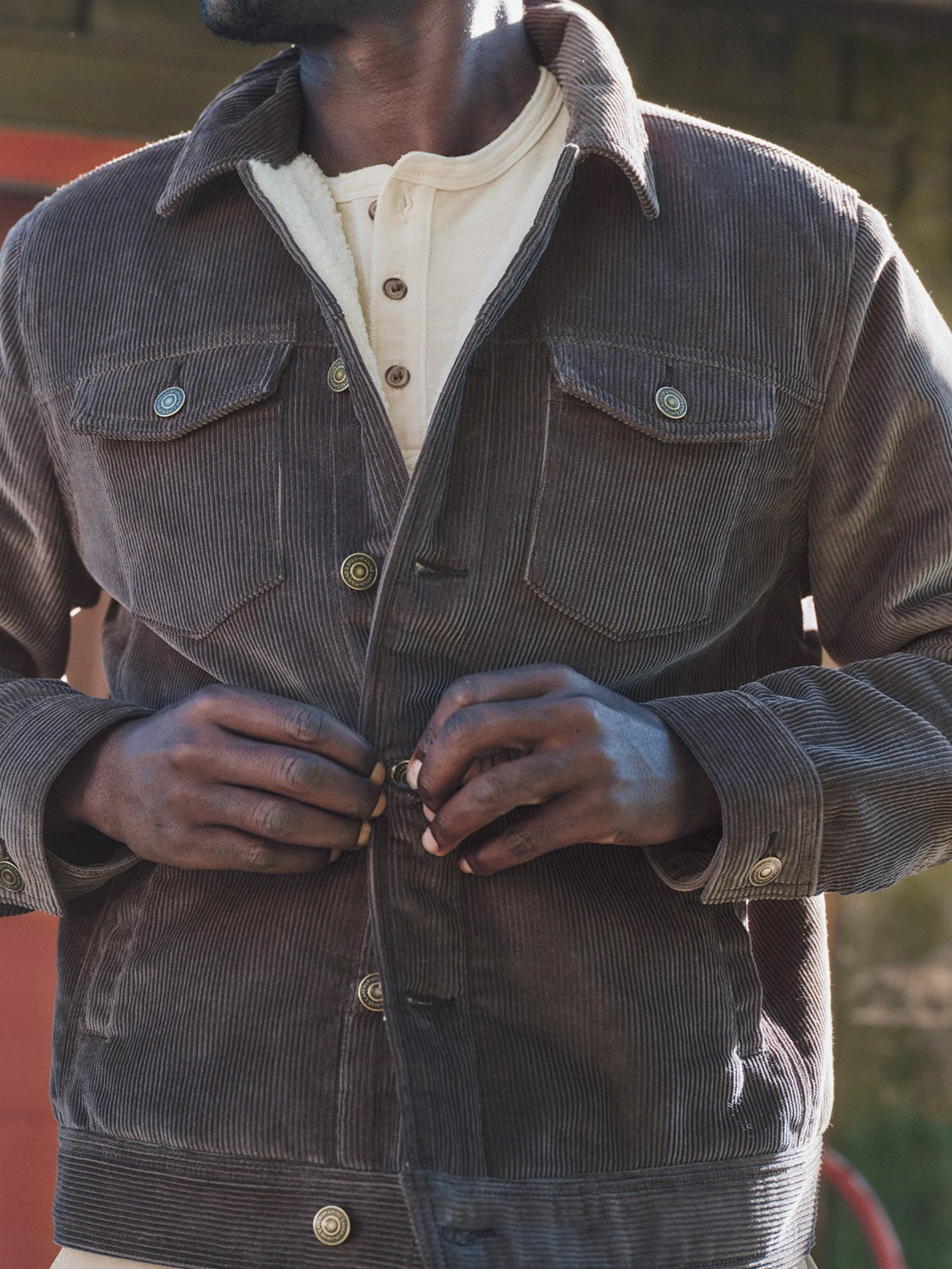 A person is buttoning up the Marine Layer Boise Cord Trucker, a brown corduroy jacket with a soft quilted lining, over a cream henley shirt, shown from the shoulders to waist with their face partially out of frame.