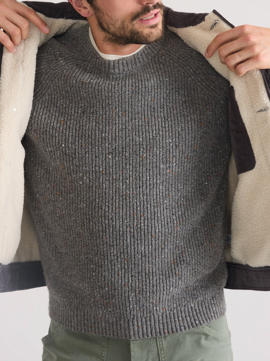 A person wearing a gray knit sweater and green pants opens the Marine Layer Boise Cord Trucker jacket with a white sherpa interior, revealing the sweater underneath. The image is cropped from shoulders to mid-thigh.