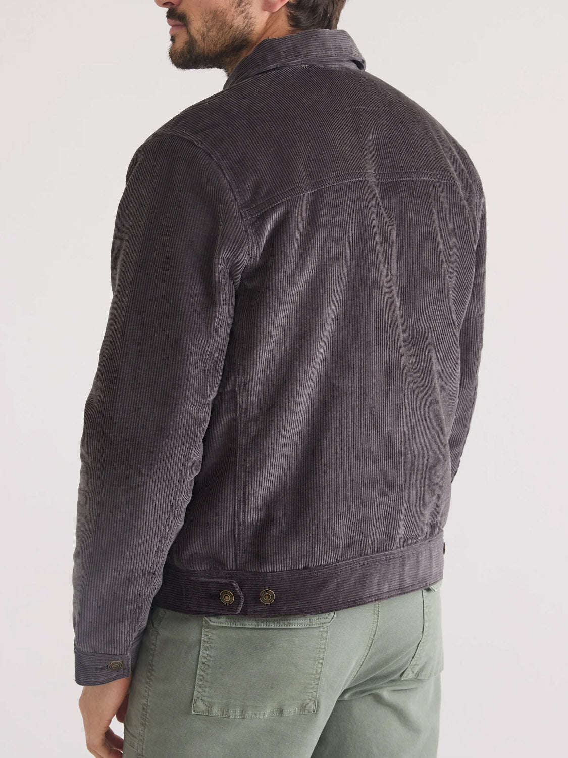 A man faces away from the camera in the Marine Layer Boise Cord Trucker jacket with a sherpa interior, paired with light green pants, standing against a plain light background.