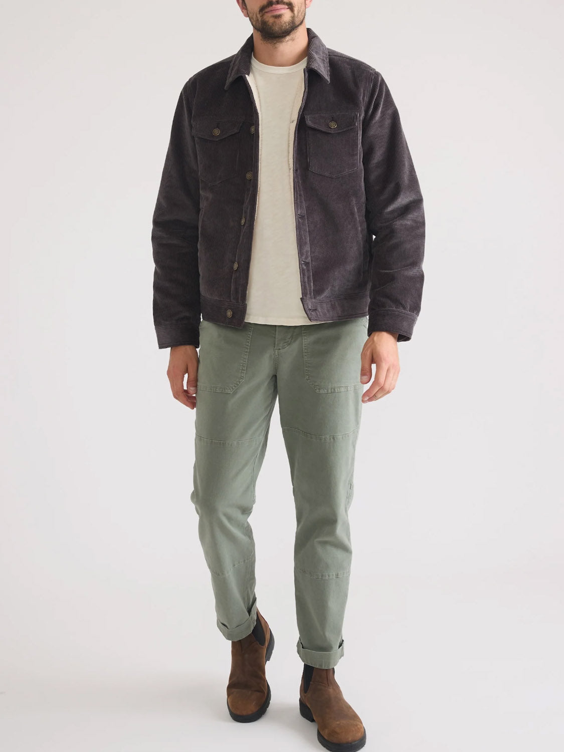 A man stands against a white background wearing the Marine Layer Boise Cord Trucker, a dark corduroy jacket with a soft quilted lining, paired with a light beige t-shirt, sage green pants, and brown suede boots. His hands are by his sides and his head slightly tilted.
