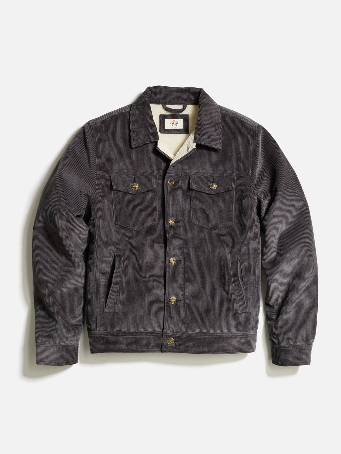 The Marine Layer Boise Cord Trucker is a dark gray corduroy jacket with a button-up front, chest and side pockets, a collar, and a soft quilted lining for added comfort and warmth.