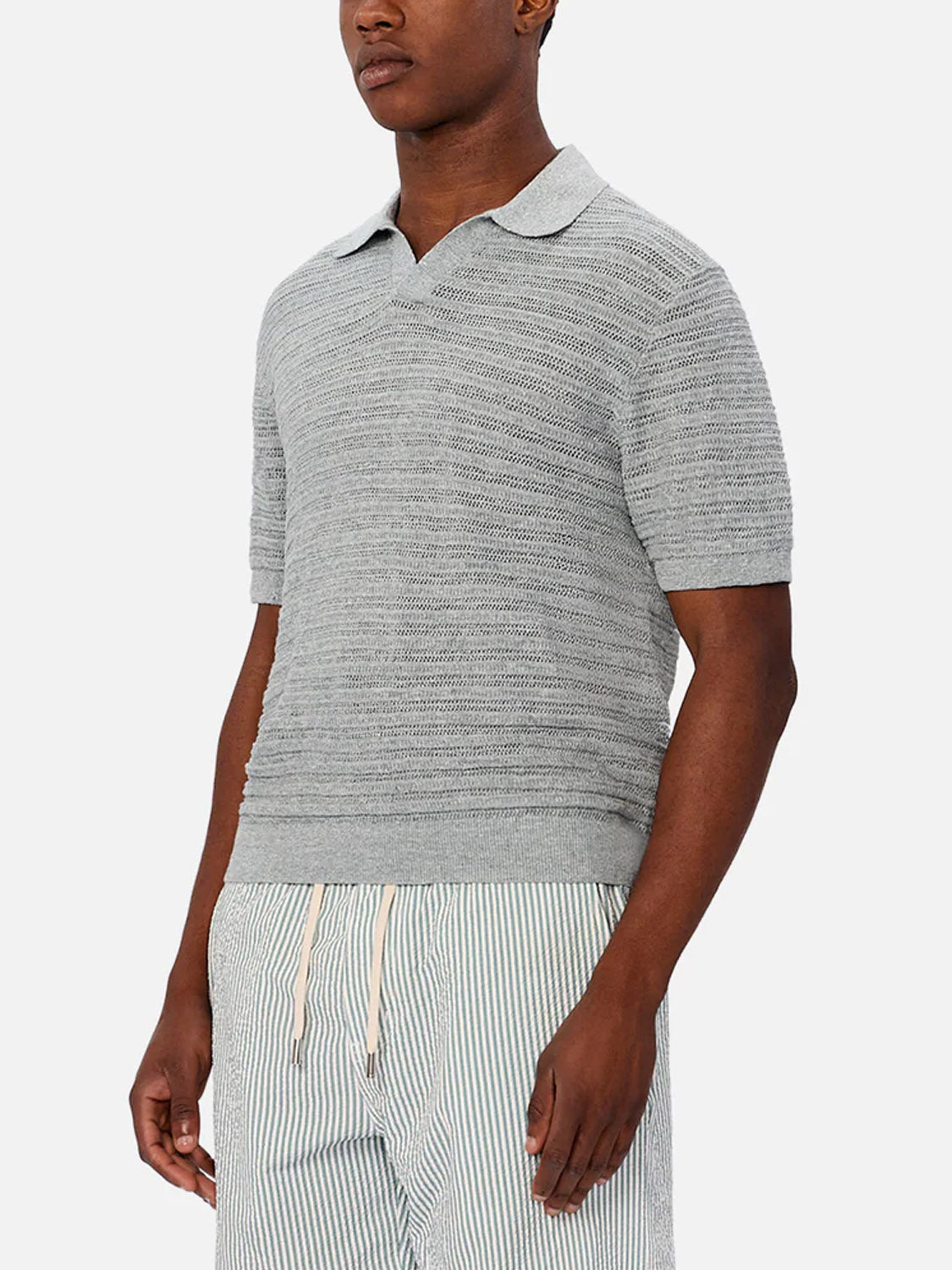 A man wearing Industrie’s The Bologna Polo in light gray stands against a plain white background, paired with light blue and white striped drawstring shorts.