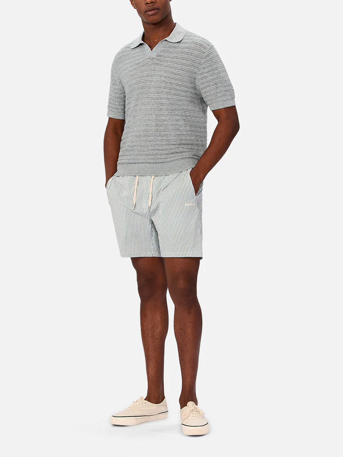 A man stands with his hands in his pockets wearing Industrie’s The Bologna Polo in light gray, paired with light blue and white striped drawstring shorts and beige sneakers, set against a plain white background.