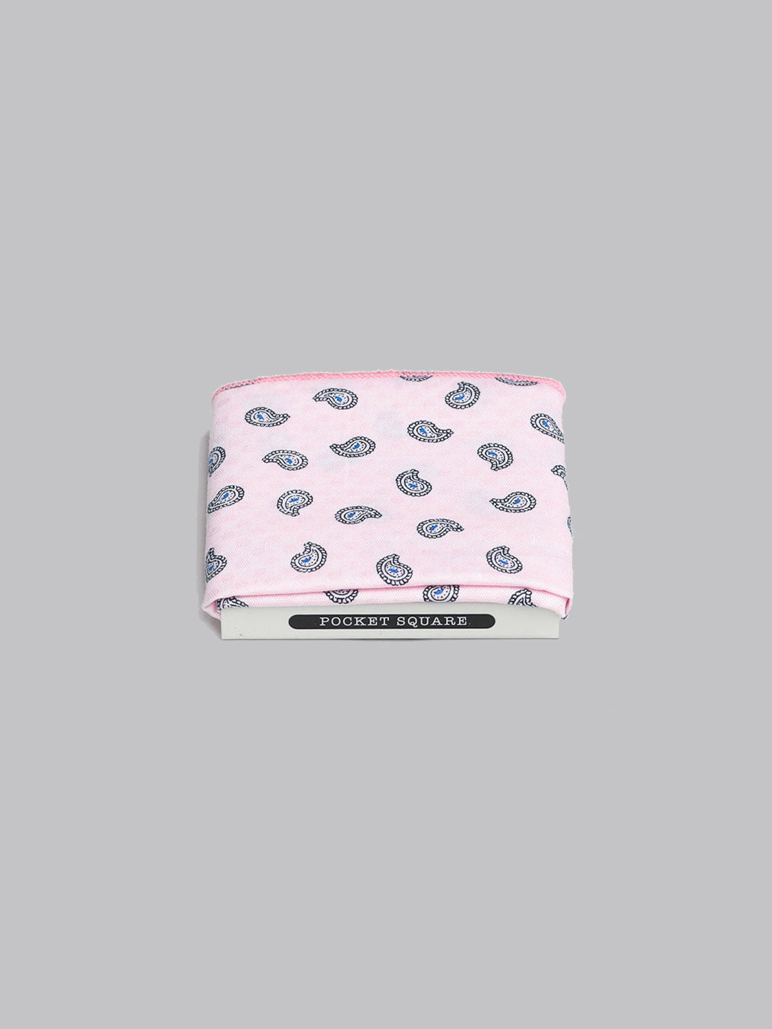 The Bombay Pocket Square by ID Menswear, handmade in Brooklyn from 100% cotton, features a folded pink design with a blue paisley pattern. It’s shown on a gray background, partially wrapped in packaging labeled POCKET SQUARE.