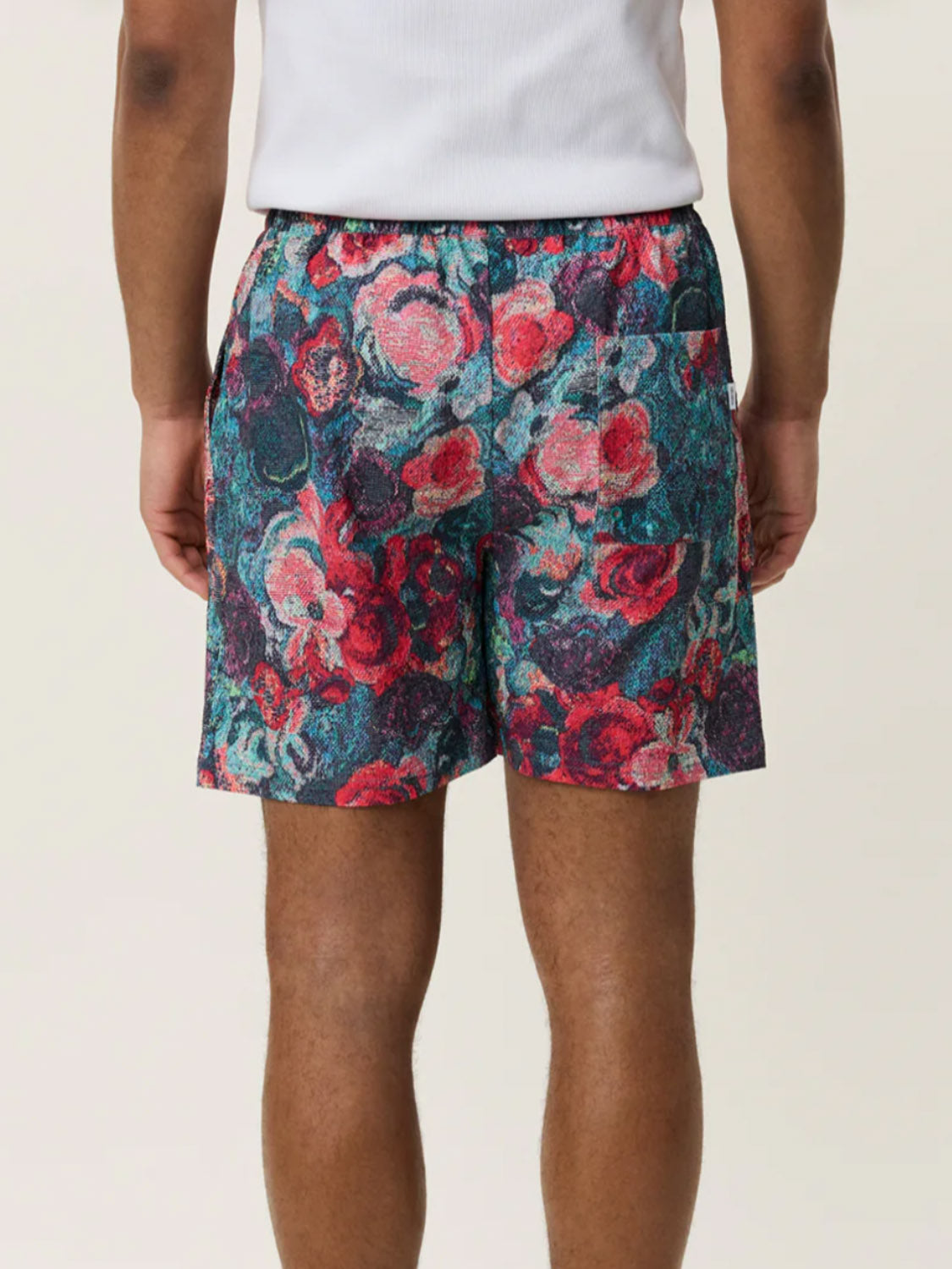 A person is seen from the back wearing Les Deux Bouquet AOP Swim Shorts with a rose print and a white top, standing against a plain light background.