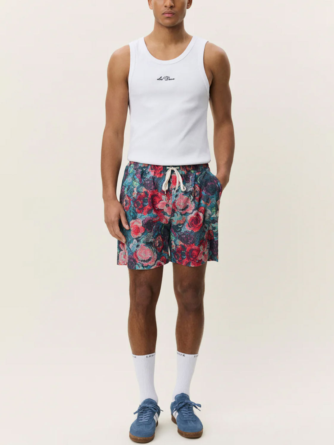 A person wearing a white tank top, Les Deux Bouquet AOP Swim Shorts with a colorful floral design, white crew socks, and blue sneakers stands against a plain light background.