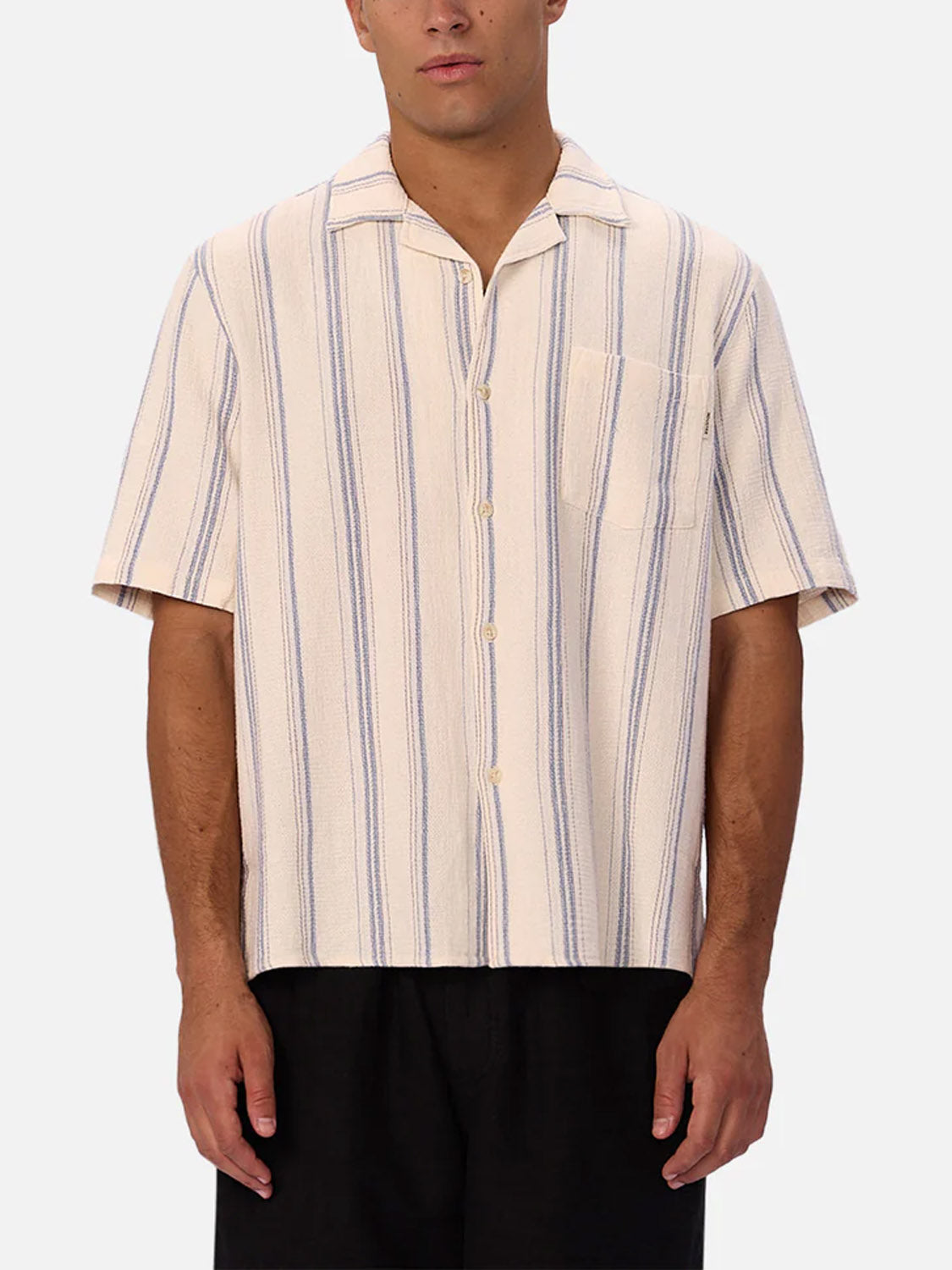 A person wearing the Industrie Bowment Short Sleeve Shirt, featuring breathable cotton, a camp collar, vertical blue and beige stripes, and a chest pocket, paired with black pants against a plain light background.