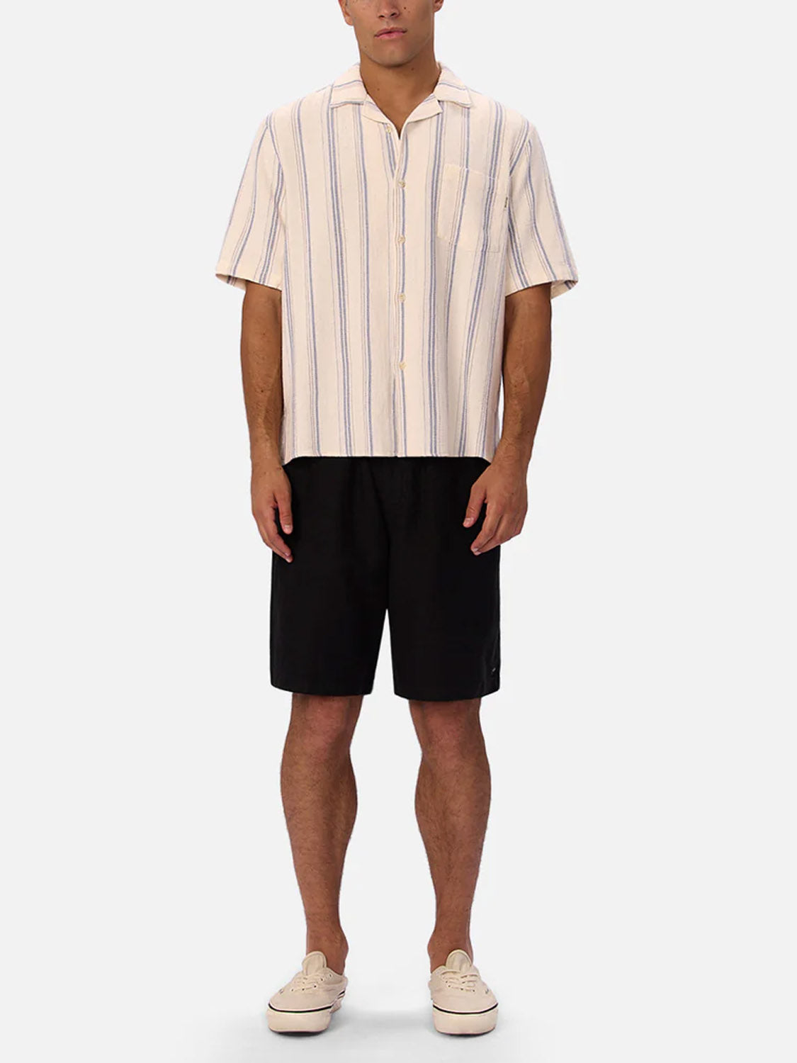 A person stands facing forward in Industrie’s Bowment Short Sleeve Shirt, featuring blue vertical stripes on cream cotton, paired with black shorts and beige sneakers. The breathable shirt pops against a plain white background.