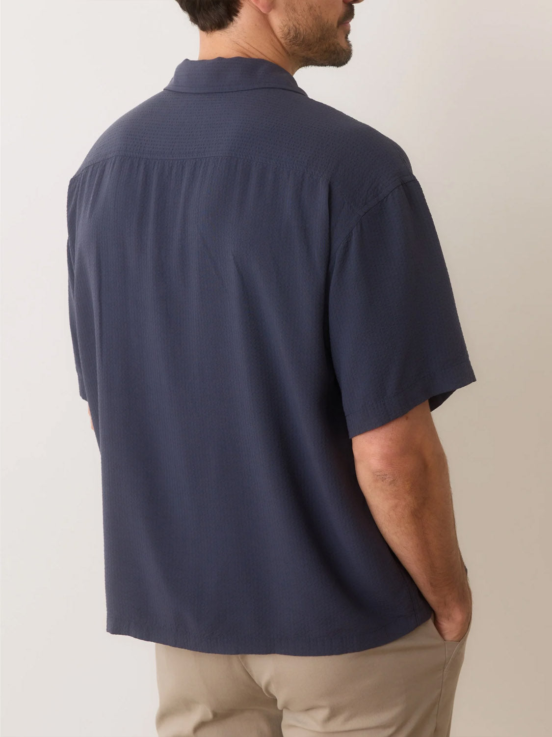 A man with hands in pockets wears the dark Marine Layer Boxy Resort Shirt crafted from TENCEL™ Lyocell, and light pants. His back faces the camera against a neutral-toned background, highlighting the cool fabric.