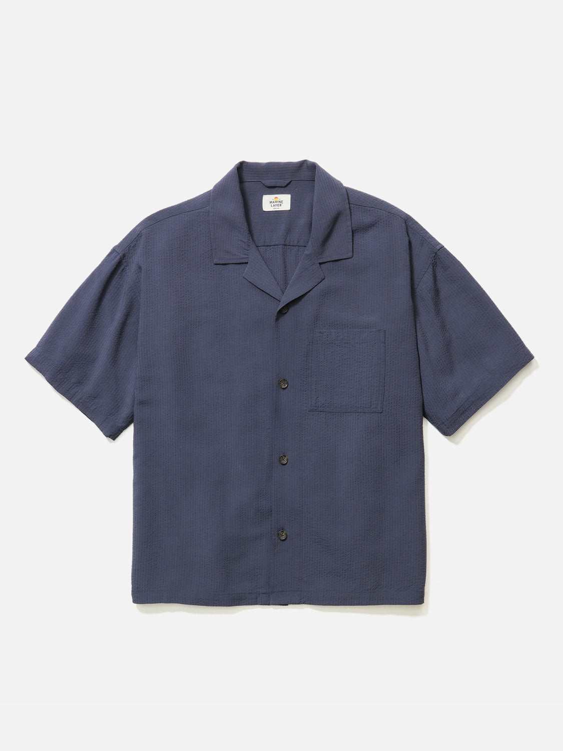 The Marine Layer Boxy Resort Shirt is a navy, short-sleeve shirt with a textured, cool-handed fabric. Featuring a resort collar, a left front pocket, and four dark buttons down the front, it is set against a plain white background, highlighting its refined yet relaxed style.