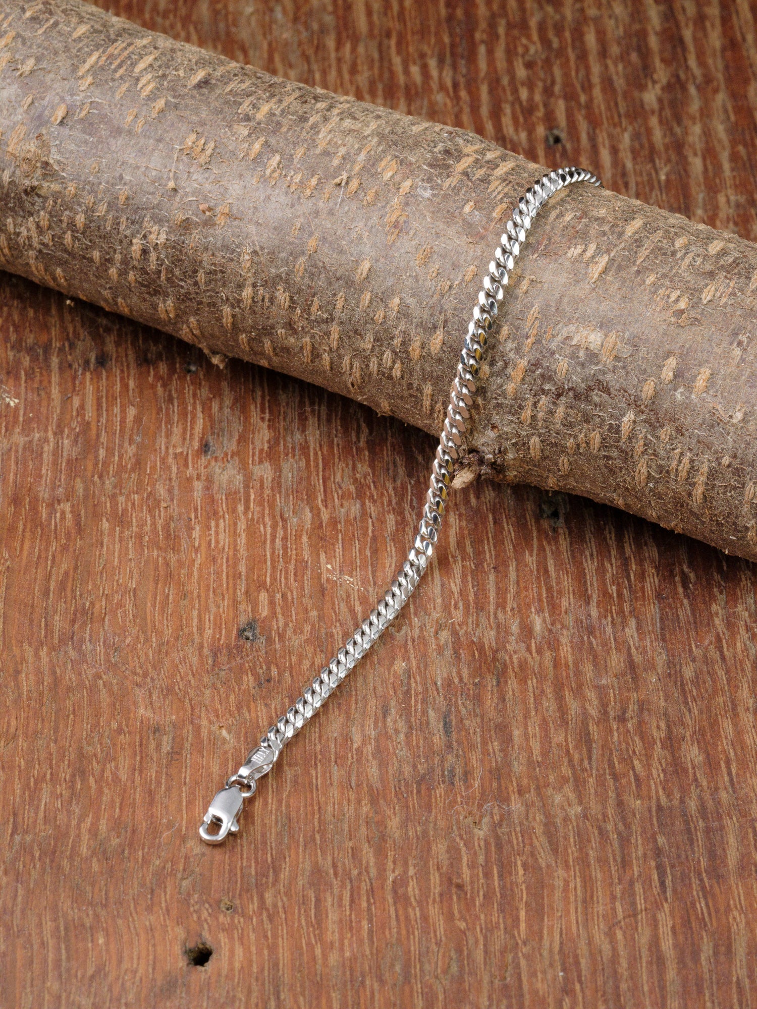 The Blaze 100 Miami Cuban Bracelet by ID Menswear, crafted in sterling silver with a lobster clasp, is shown draped over a cylindrical wooden piece on a textured brown wood surface.