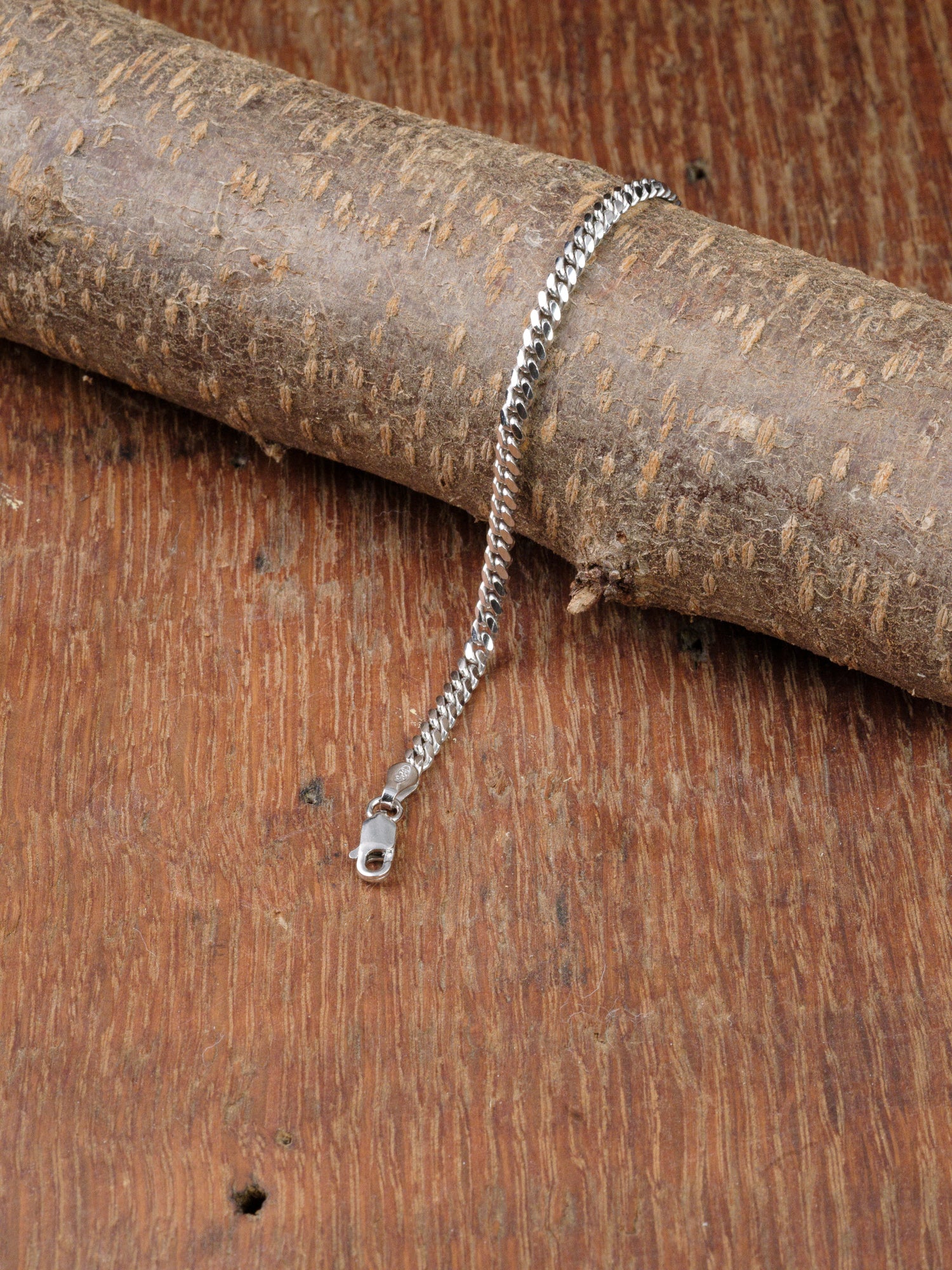 The Blaze 100 Miami Cuban Bracelet by ID Menswear, crafted in sterling silver, is displayed with a clasp on a textured brown wood surface. Its classic Miami Cuban chain curves elegantly atop a rough cylindrical wooden piece.