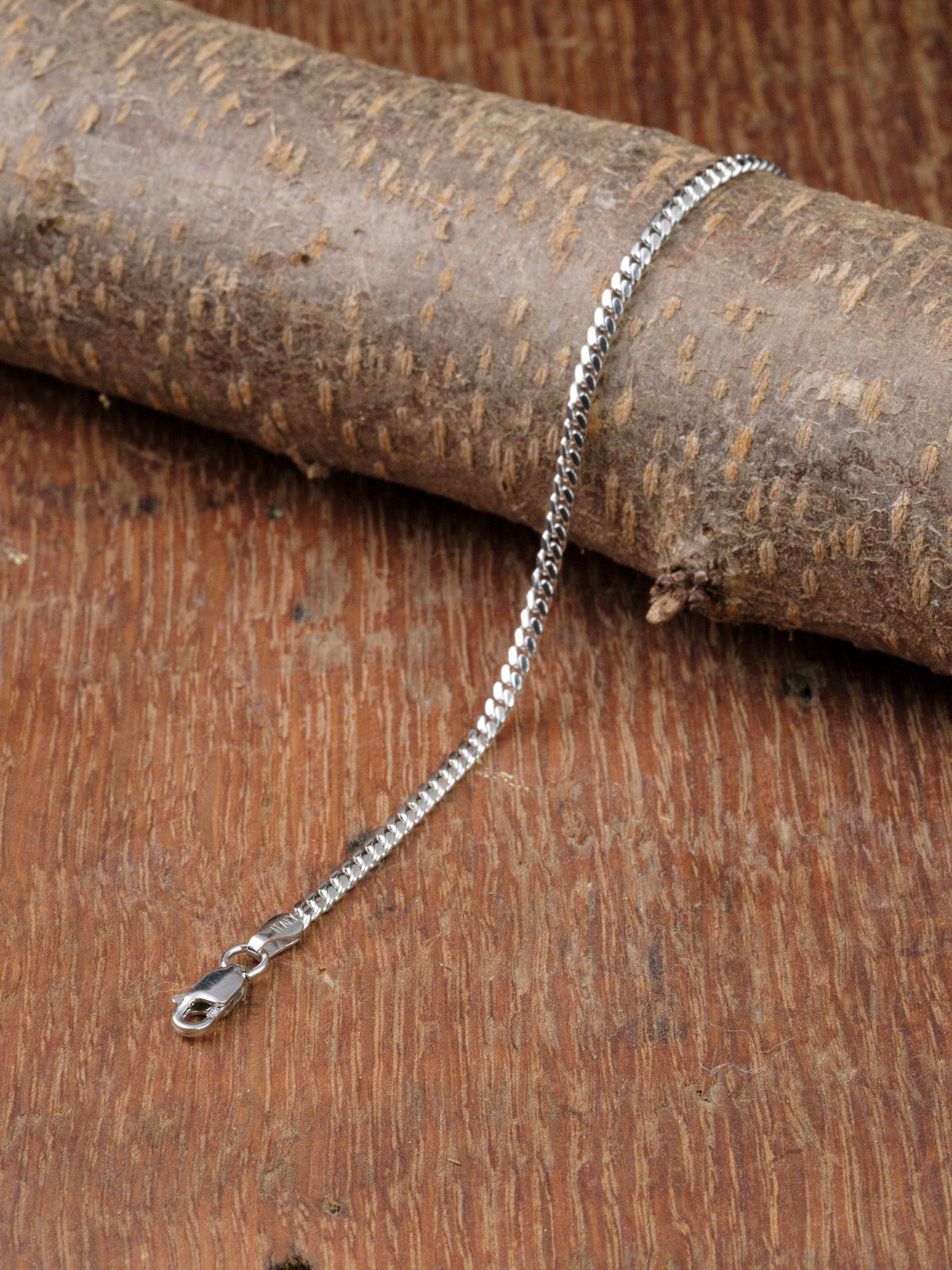 The Blaze 80 Miami Cuban Bracelet by ID Menswear, a sleek silver chain with a lobster clasp, is showcased draped elegantly over a textured wooden surface.