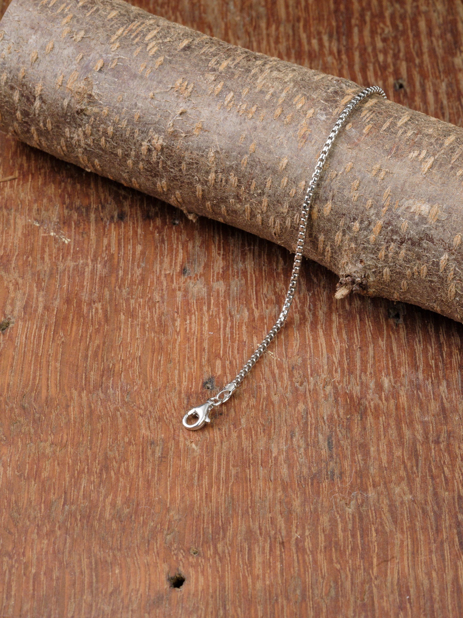 The Ezra 35 Bracelet by ID Menswear, featuring a sterling silver box chain with a lobster clasp, is elegantly displayed on a textured wooden surface and draped over tree bark.