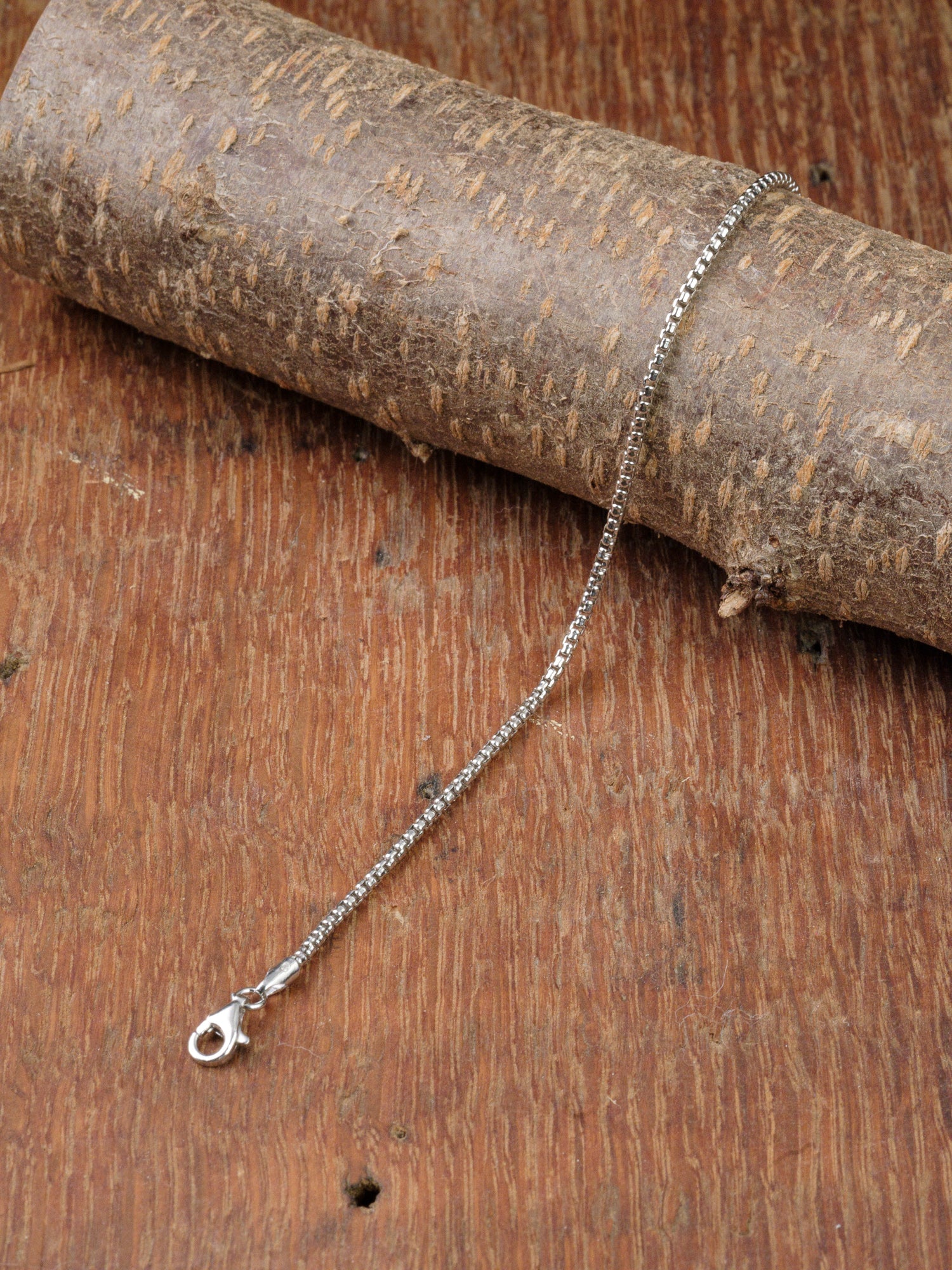 The Ezra 35 Bracelet by ID Menswear, featuring a sterling silver box chain and lobster clasp, is displayed on a rustic wooden surface, partially draped over a cylindrical wooden piece.