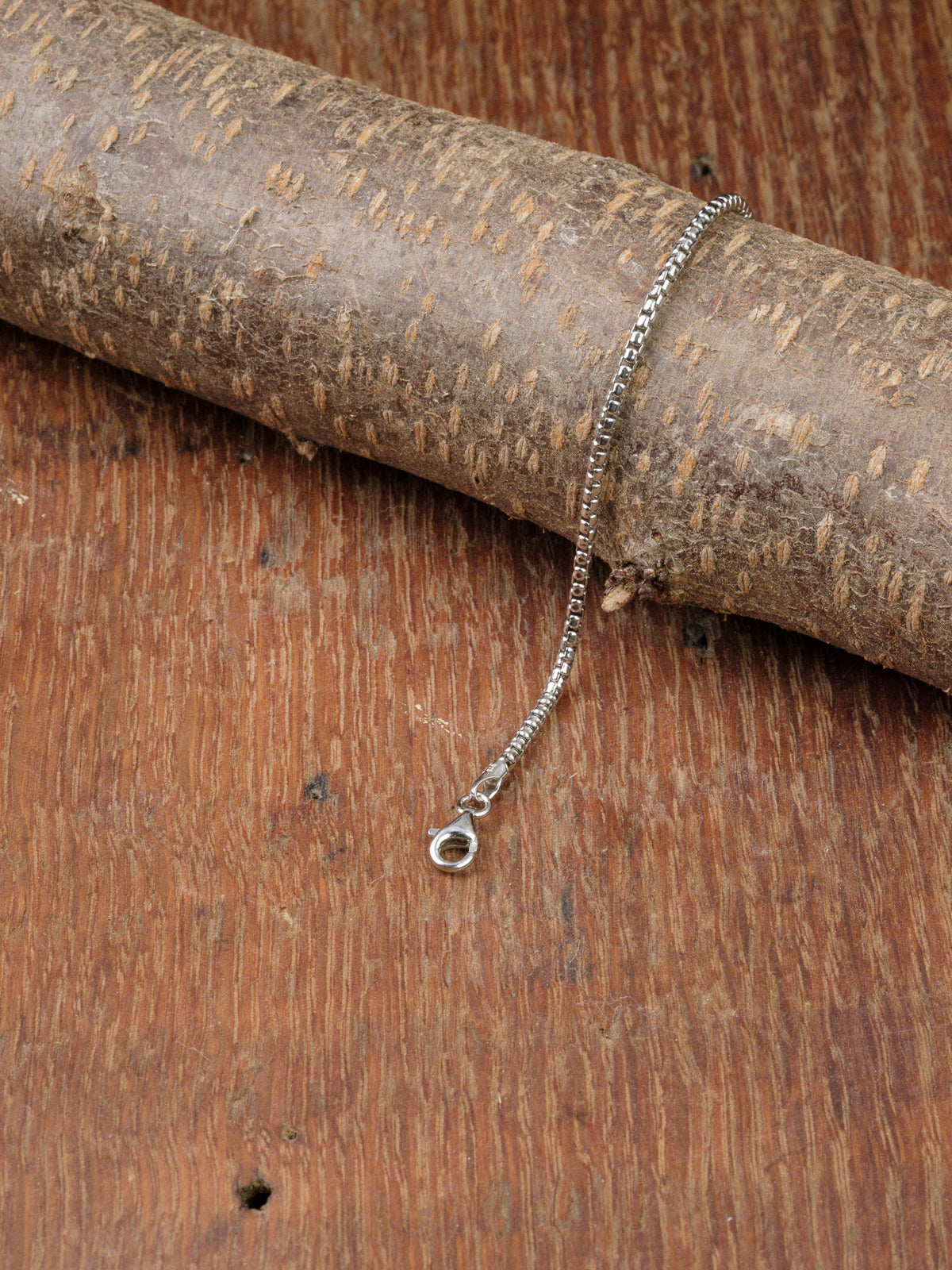 The Ezra 40 Bracelet by ID Menswear, featuring a sterling silver box chain and clasp, is displayed draped over tree bark on a wooden surface.