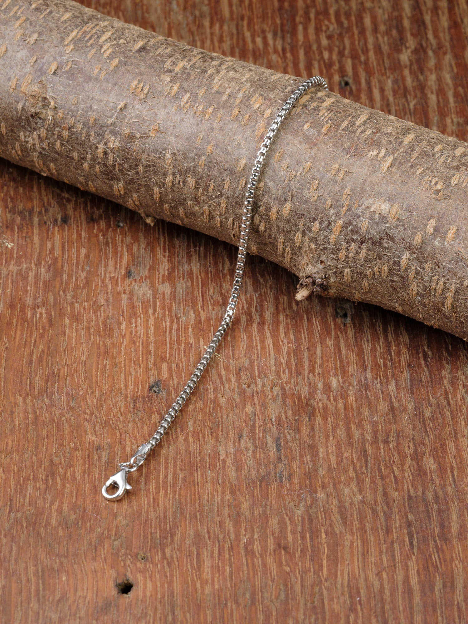 The Ezra 40 Bracelet by ID Menswear, crafted in Italy from sterling silver with a box chain and lobster clasp, is displayed on a textured wooden surface draped over rustic wood, highlighting its timeless elegance and expert craftsmanship.