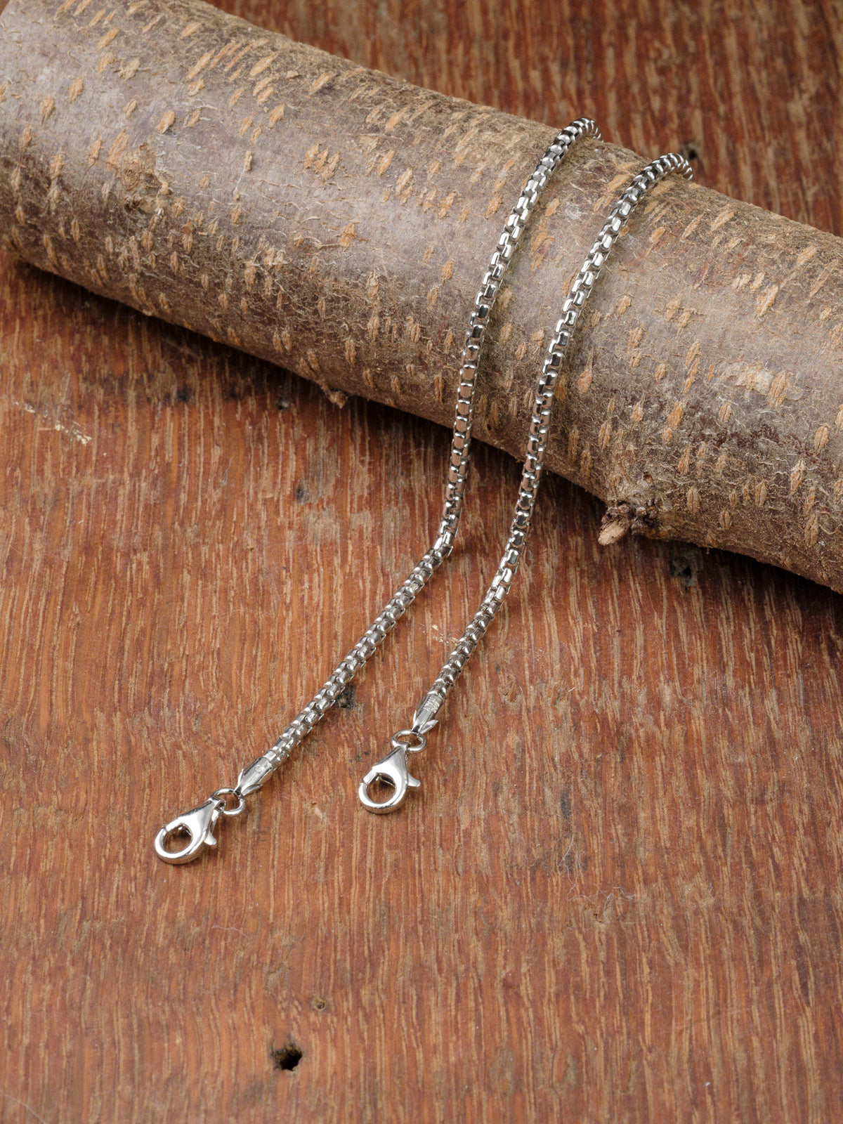 A silver chain necklace with a lobster clasp is laid on wood, draped over a textured piece, next to the sleek Ezra 50 Bracelet by ID Menswear in 925 Sterling Silver for an elegant pairing.