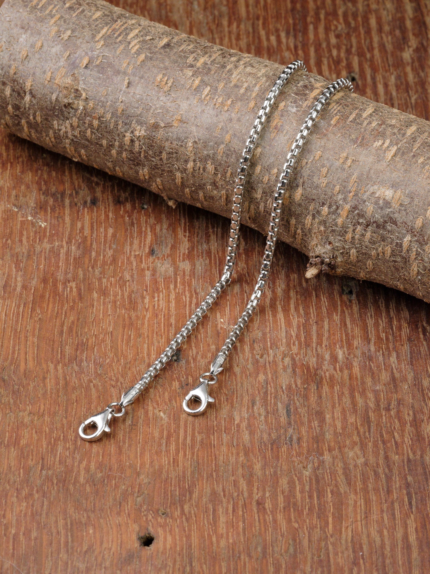 A silver chain necklace with a lobster clasp is laid on wood, draped over a textured piece, next to the sleek Ezra 50 Bracelet by ID Menswear in 925 Sterling Silver for an elegant pairing.
